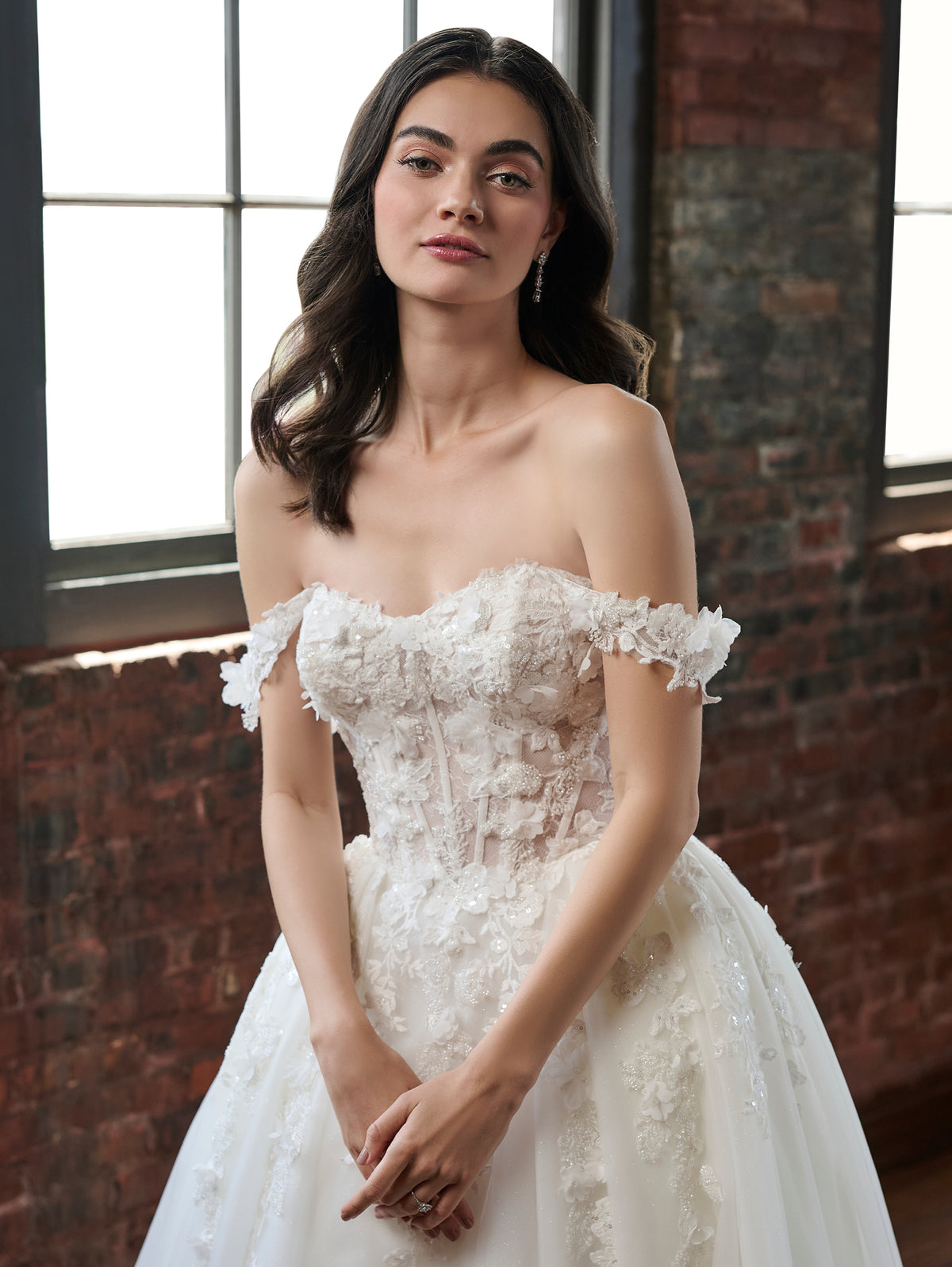 A woman wearing an off-the-shoulder white lace wedding gown with a fitted bodice and full tulle skirt, standing indoors against a brick wall and window. / Off the shoulder ballgown with a textured embellishment created with over lace, lace motifs & beaded 3D petals.