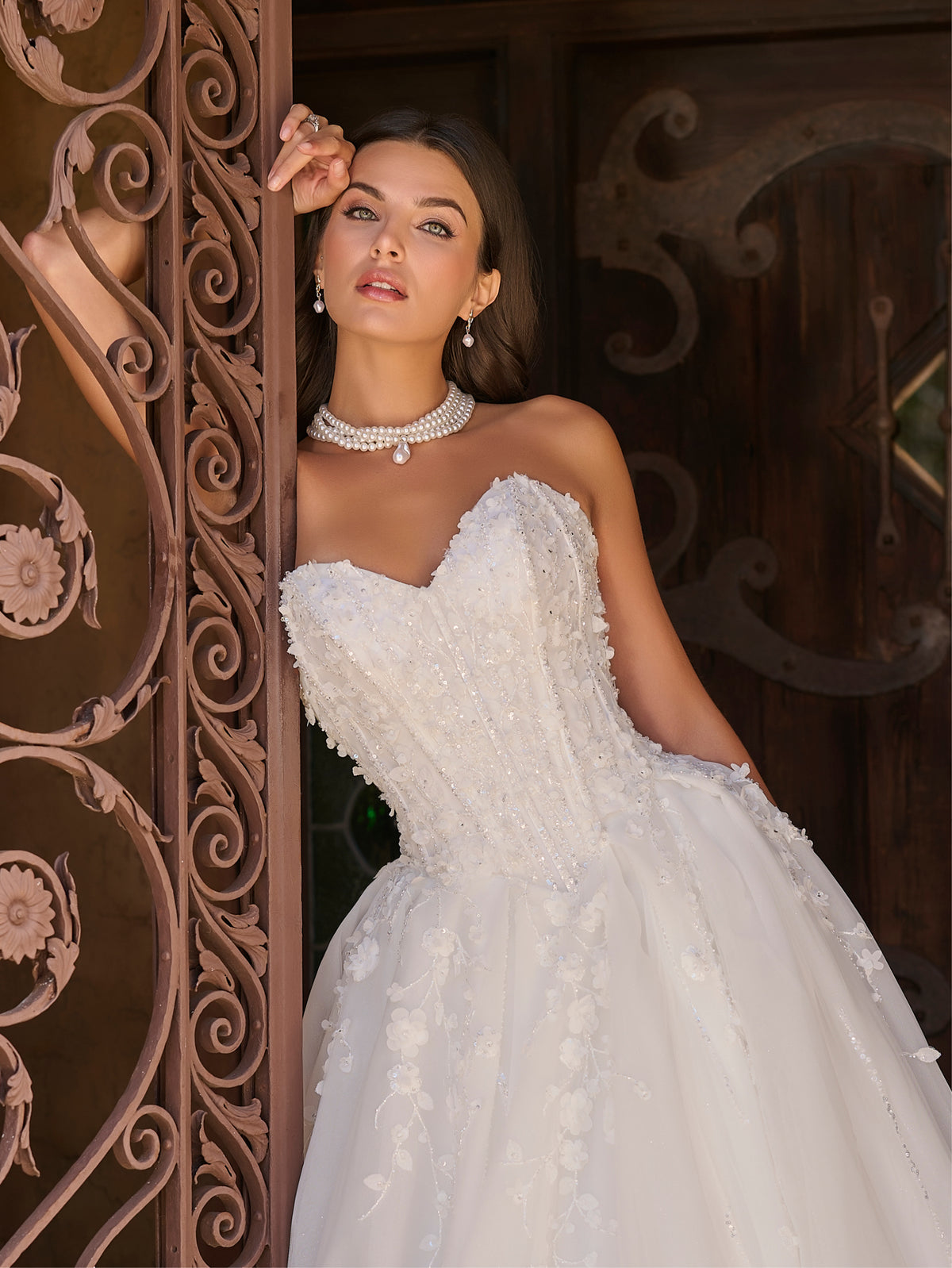 Woman in a white wedding dress standing behind an ornate wooden door. / Ball gown with sweetheart neckline and exposed boning decorated with hand beading and 3D lace motifs. Lace up back closure.