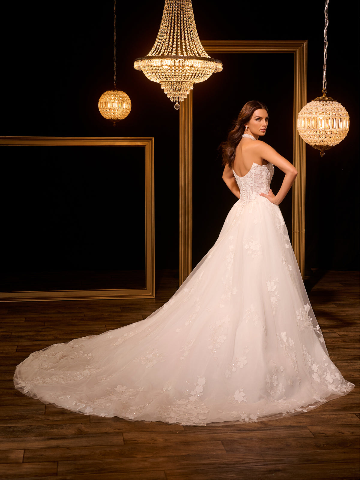 Woman in a white wedding dress standing in a room with chandeliers. / Ball gown with floral lace motifs and split on side skirt.