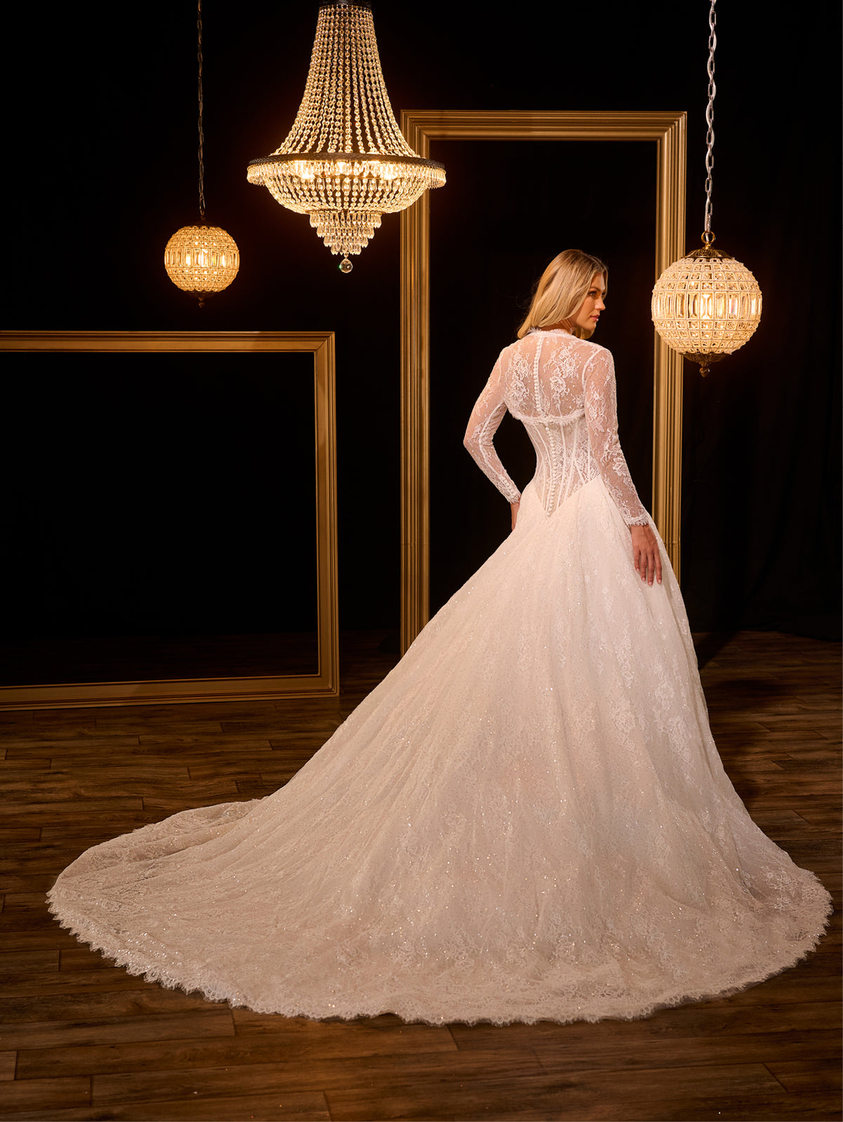 Woman in a white lace wedding dress standing in a room with chandeliers. / Ball gown in all over Chantilly lace with detachable lace bolero. Scooped neckline, buttons/zipper back closure.