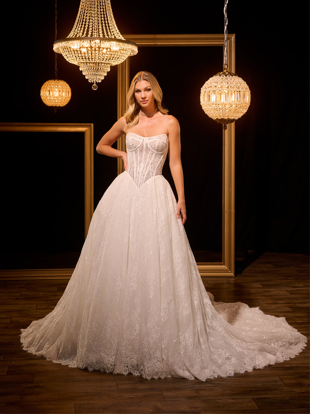 Woman in a white wedding dress standing in a dimly lit room with chandeliers. / Ball gown in all over Chantilly lace with detachable lace bolero. Scooped neckline, buttons/zipper back closure.