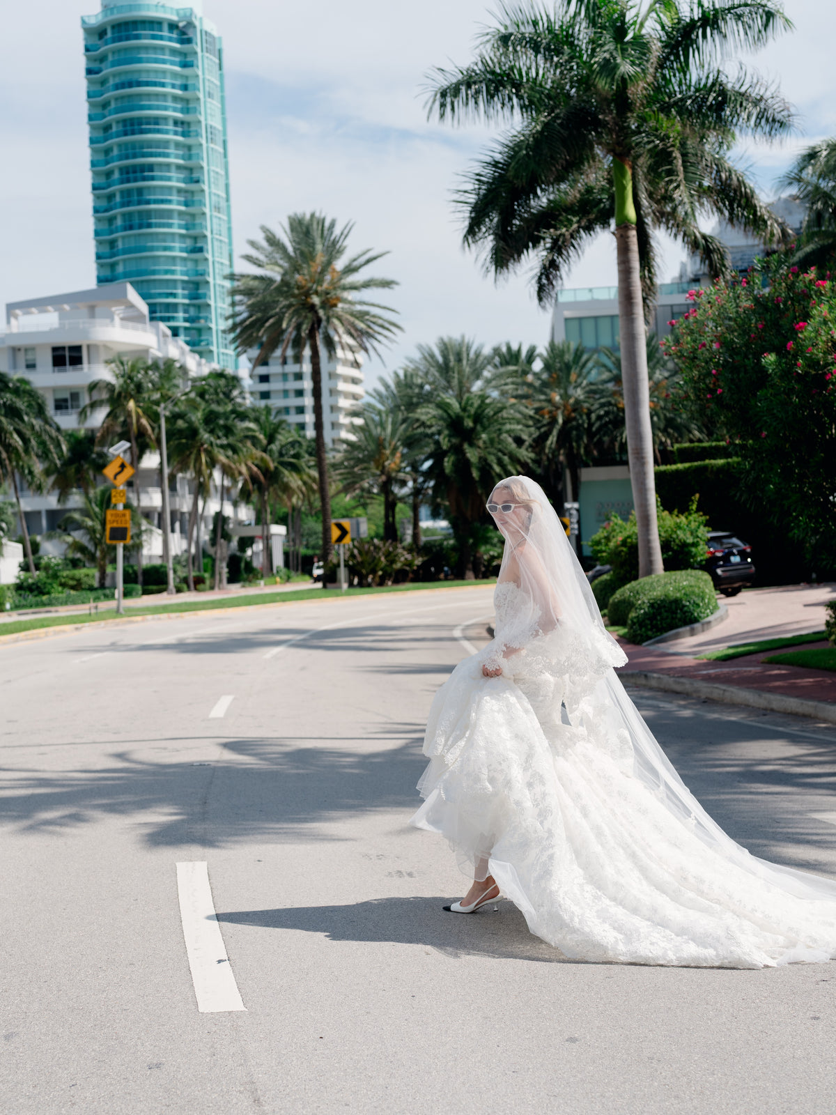 A bride in a voluminous white lace wedding gown and long veil walks across a city street lined with palm trees and modern buildings under a bright sky. / Mermaid style in all over lace with scooped neckline and lace up back.