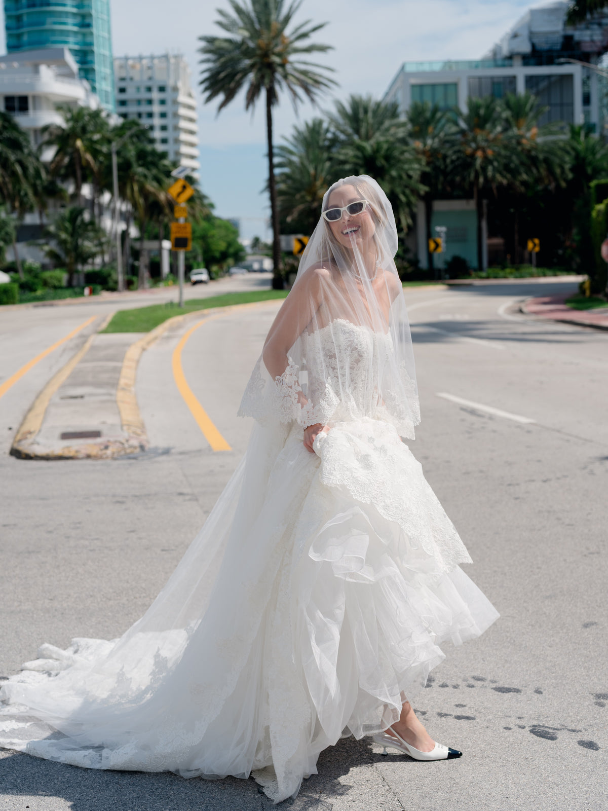 A woman wearing a white strapless wedding gown with a long sheer veil and white pointed flats stands on an urban street with palm trees and buildings in the background. / Mermaid style in all over lace with scooped neckline and lace up back.