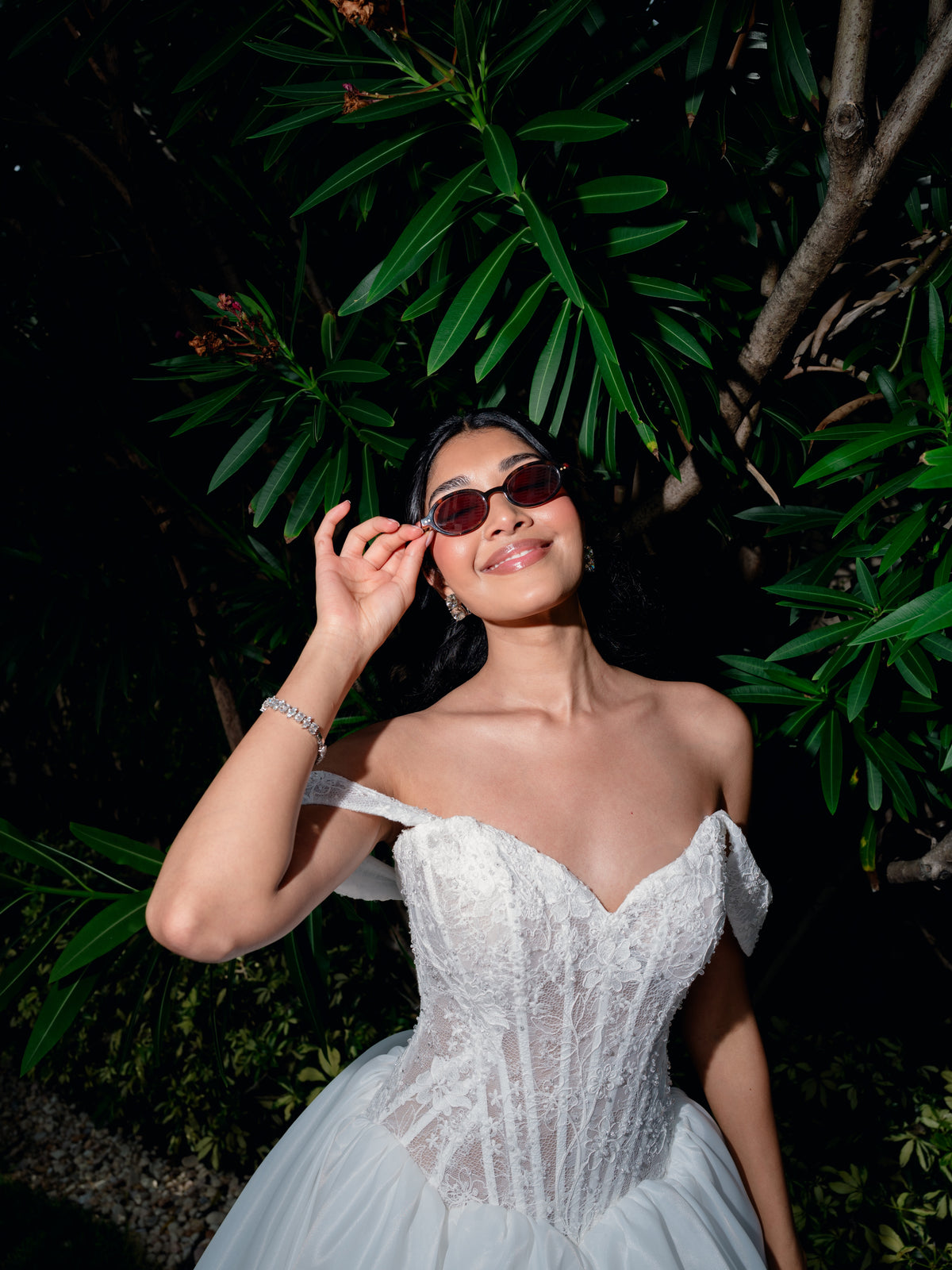 A woman wearing a white off-the-shoulder lace corset gown and dark sunglasses, posing outdoors against a dark background of green foliage. / Ball gown with semi-sheer lace bodice and organza skirt. Sweetheart neckline with off-shoulder lace straps. Lace up back closure.