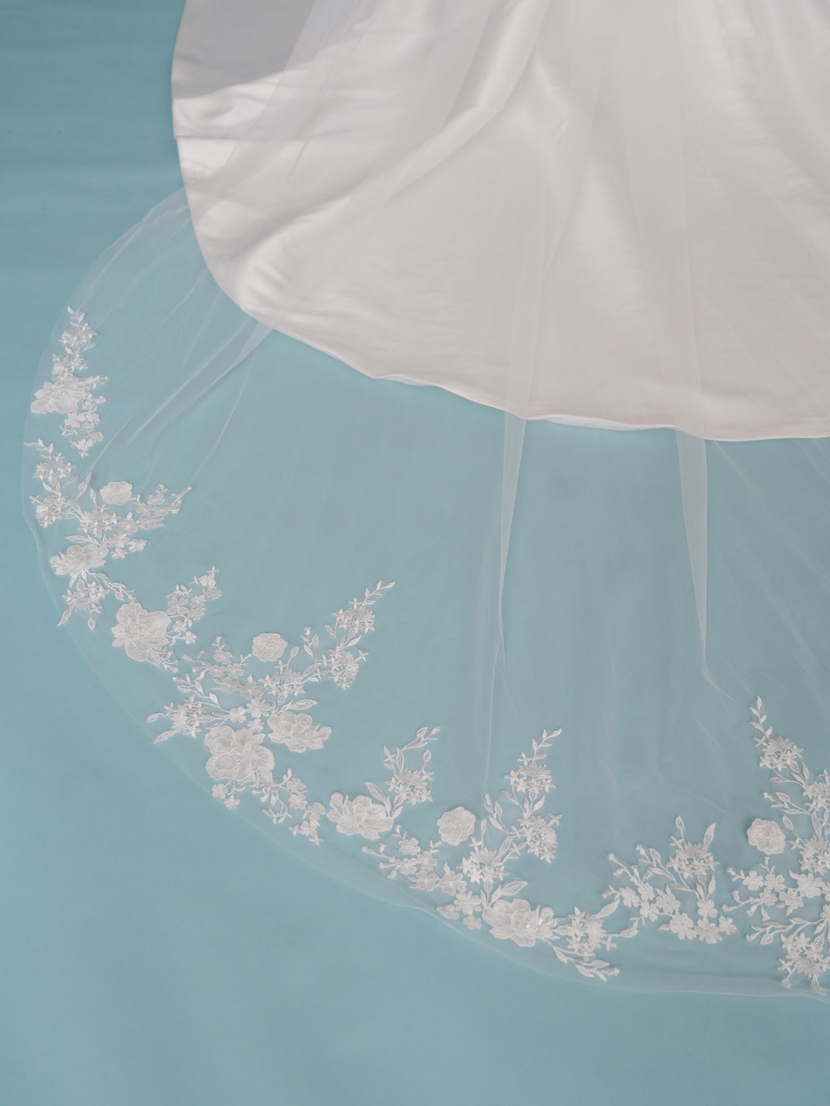 White bridal veil with delicate floral lace embroidery along the lower edge, draped over a white satin bridal veil layer on a light blue background. Long veil with lace motifs scattered around the bottom area.