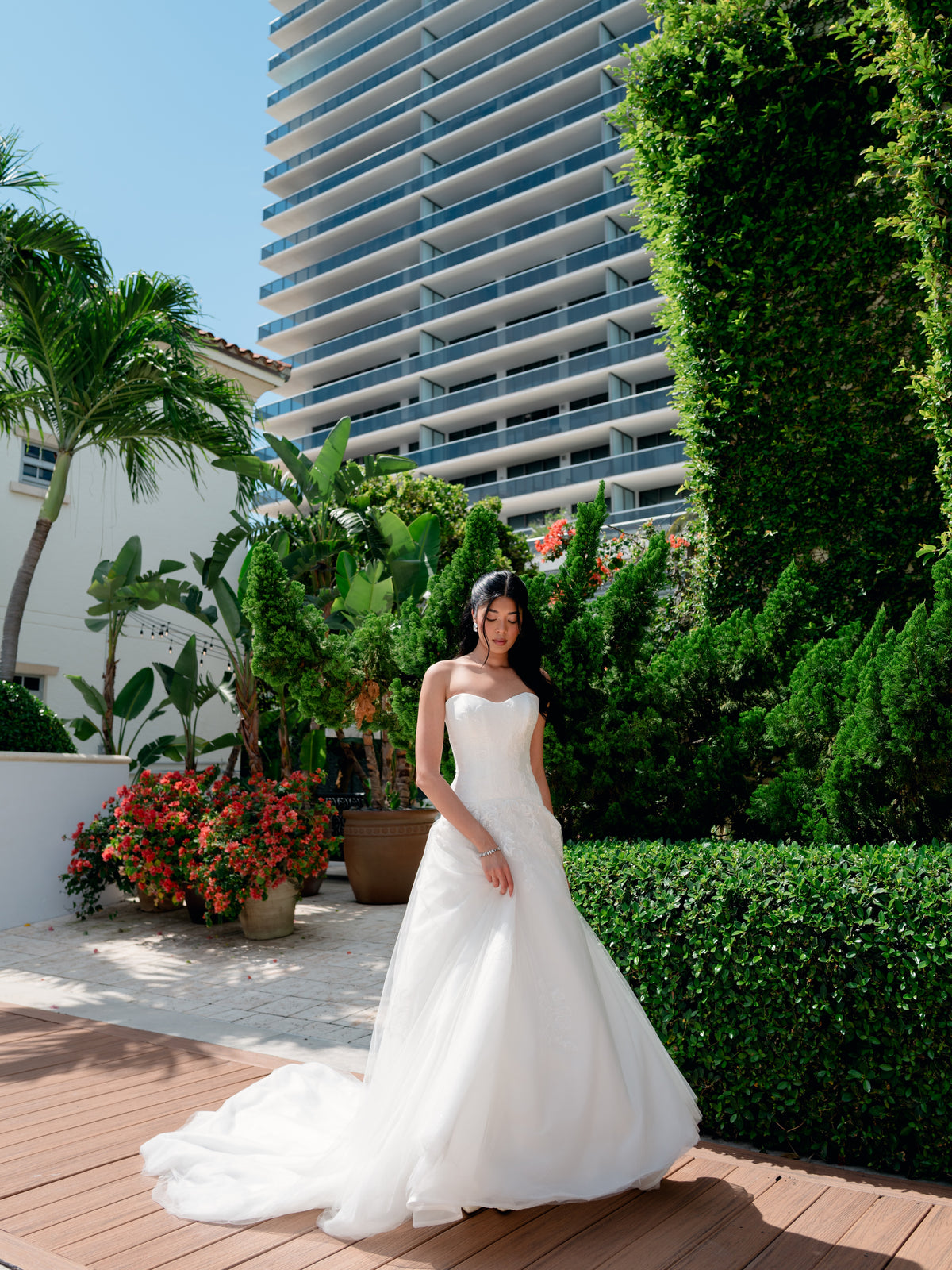 A woman wearing a strapless white wedding gown stands on a wooden deck outdoors, with lush green hedges, potted plants, and a tall modern high-rise building in the background. / Aline tulle gown with drop waist with mikado bodice and tulle skirt, decorated with shimmer lace motifs. Scooped strapless neckline.