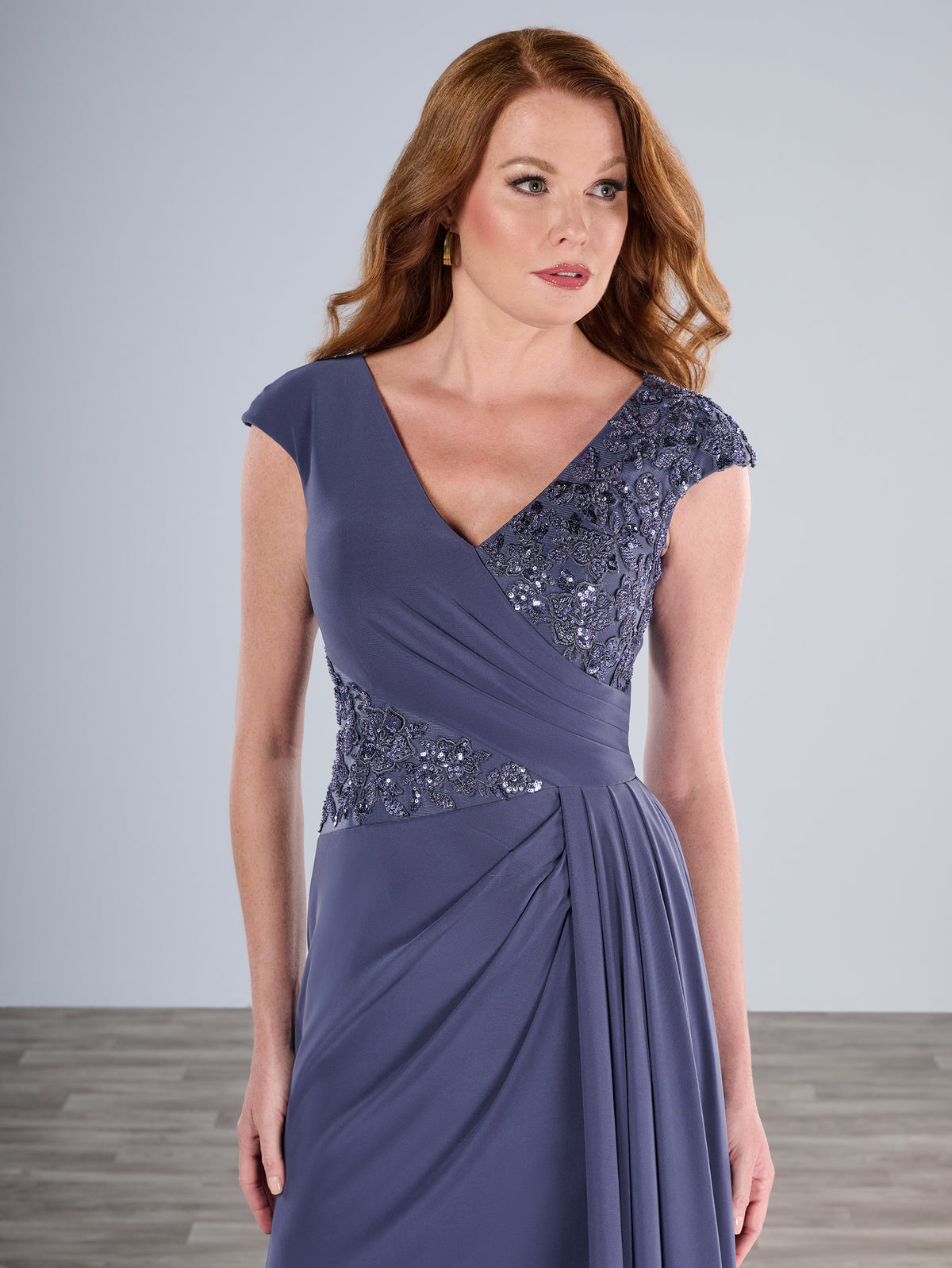 Woman wearing a purple evening gown with sequin detailing on a plain background / Slim A-line jersey gown features a combination of pleating and beaded lace. V-neck with cup sleeves. Side draping on front skirt.