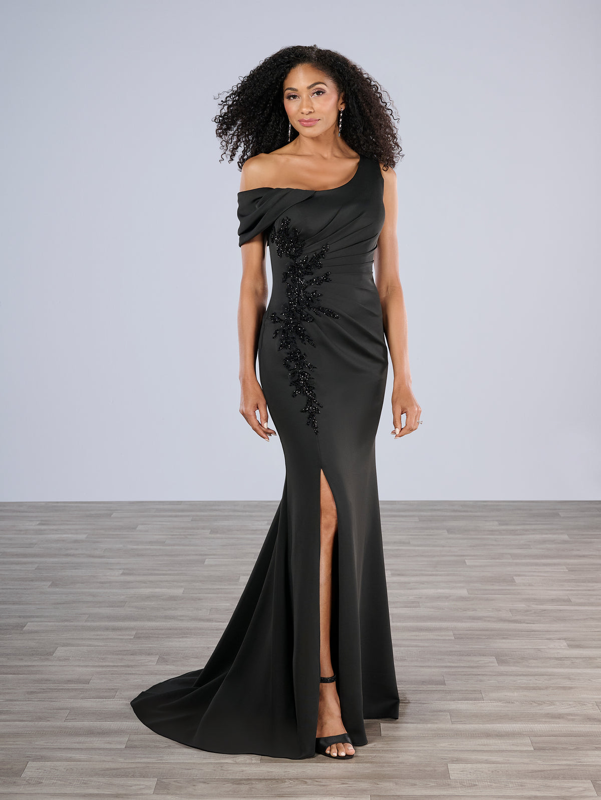 Woman wearing a black off-shoulder gown with floral details on a gray background / Slim gown with side draping and asymmetrical shoulder and off-shoulder strap on one side. The dress is decorated with beautiful, beaded motifs on the right front side and features a side slit.