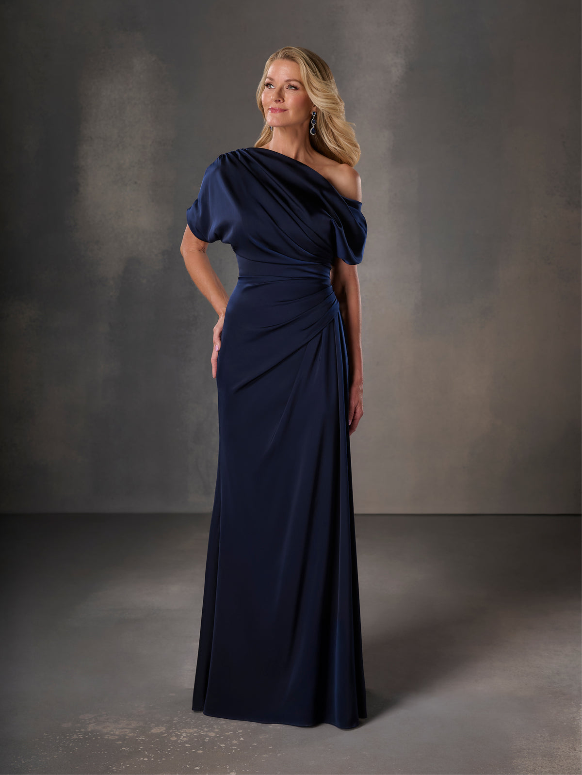 Woman wearing a navy blue evening gown against a gray background / Stretch charmeuse slim A-line with asymmetrical neckline and off-shoulder sleeve on one side.