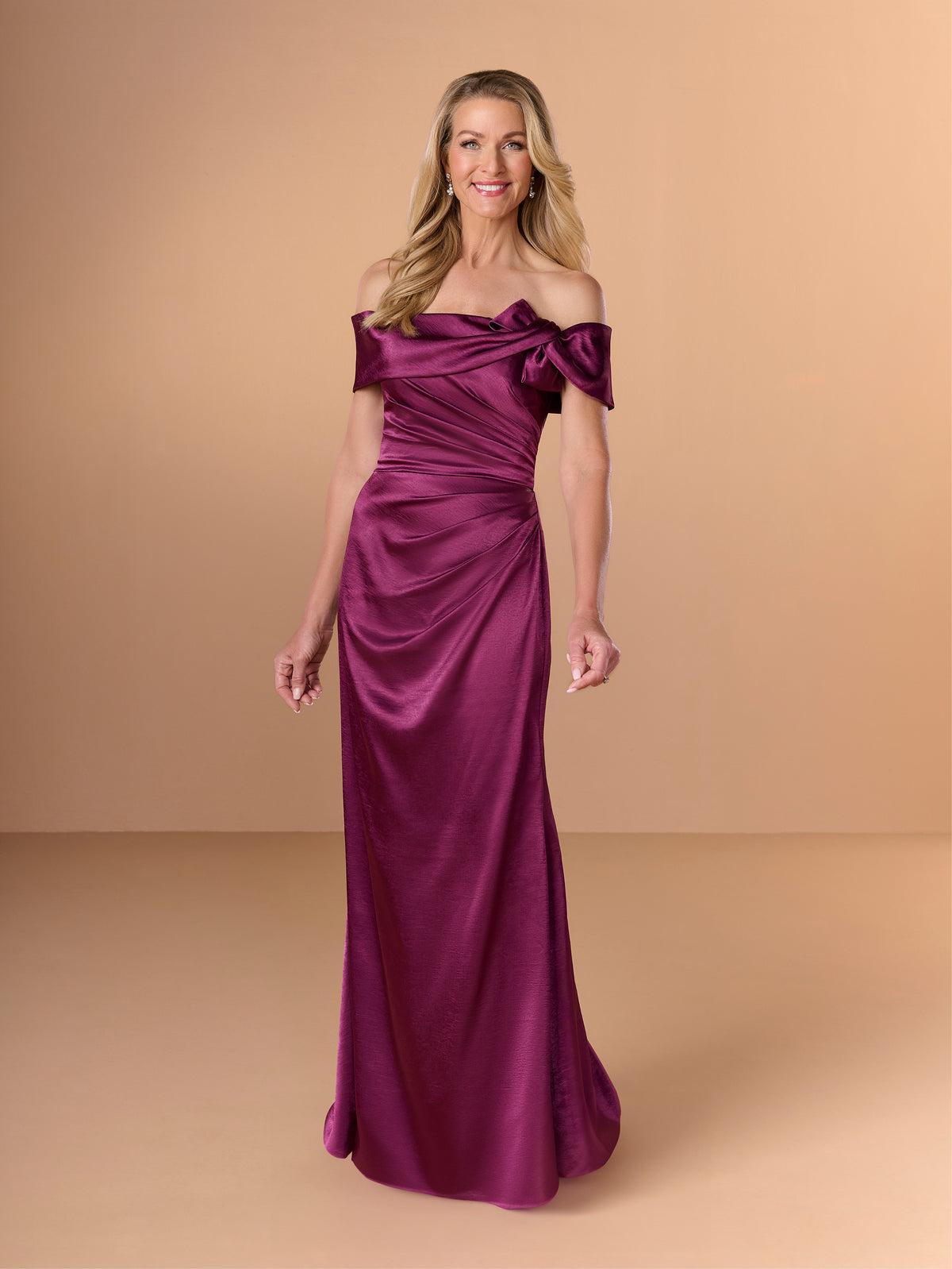 A woman modeling an off-the-shoulder floor-length gown in rich purple satin against a solid warm beige studio backdrop. / Slim charmeuse style with straight neckline and off-shoulders collar with twist and bow on side front. Pleating on front dress.