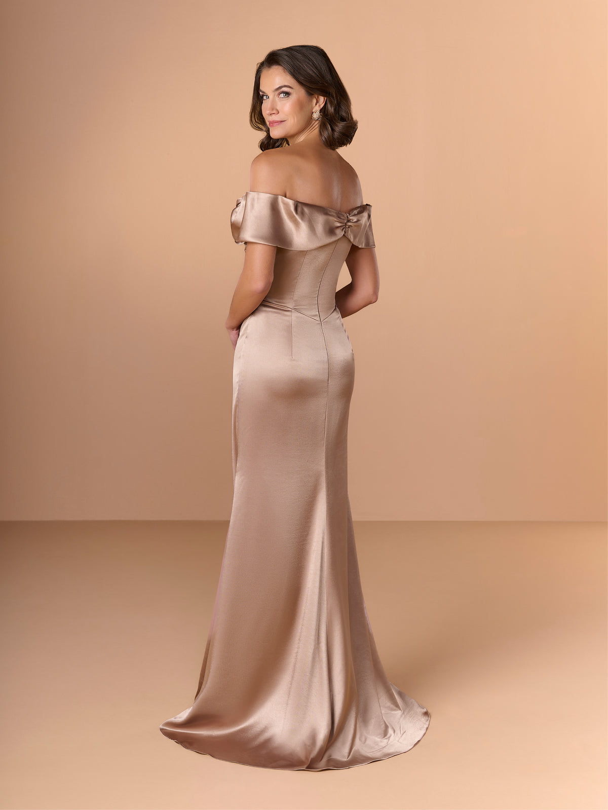 Woman wearing a satin off-shoulder gown against a beige background / Slim charmeuse style with straight neckline and off-shoulders collar with twist and bow on side front. Pleating on front dress.