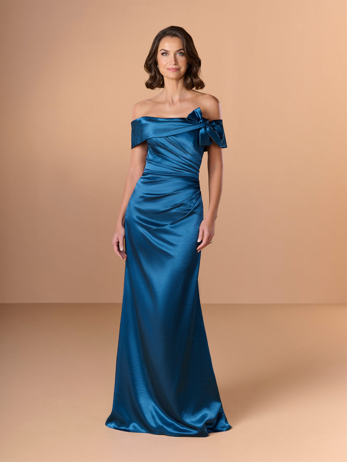 Woman wearing a blue off-shoulder gown against a beige background / Slim charmeuse style with straight neckline and off-shoulders collar with twist and bow on side front. Pleating on front dress.