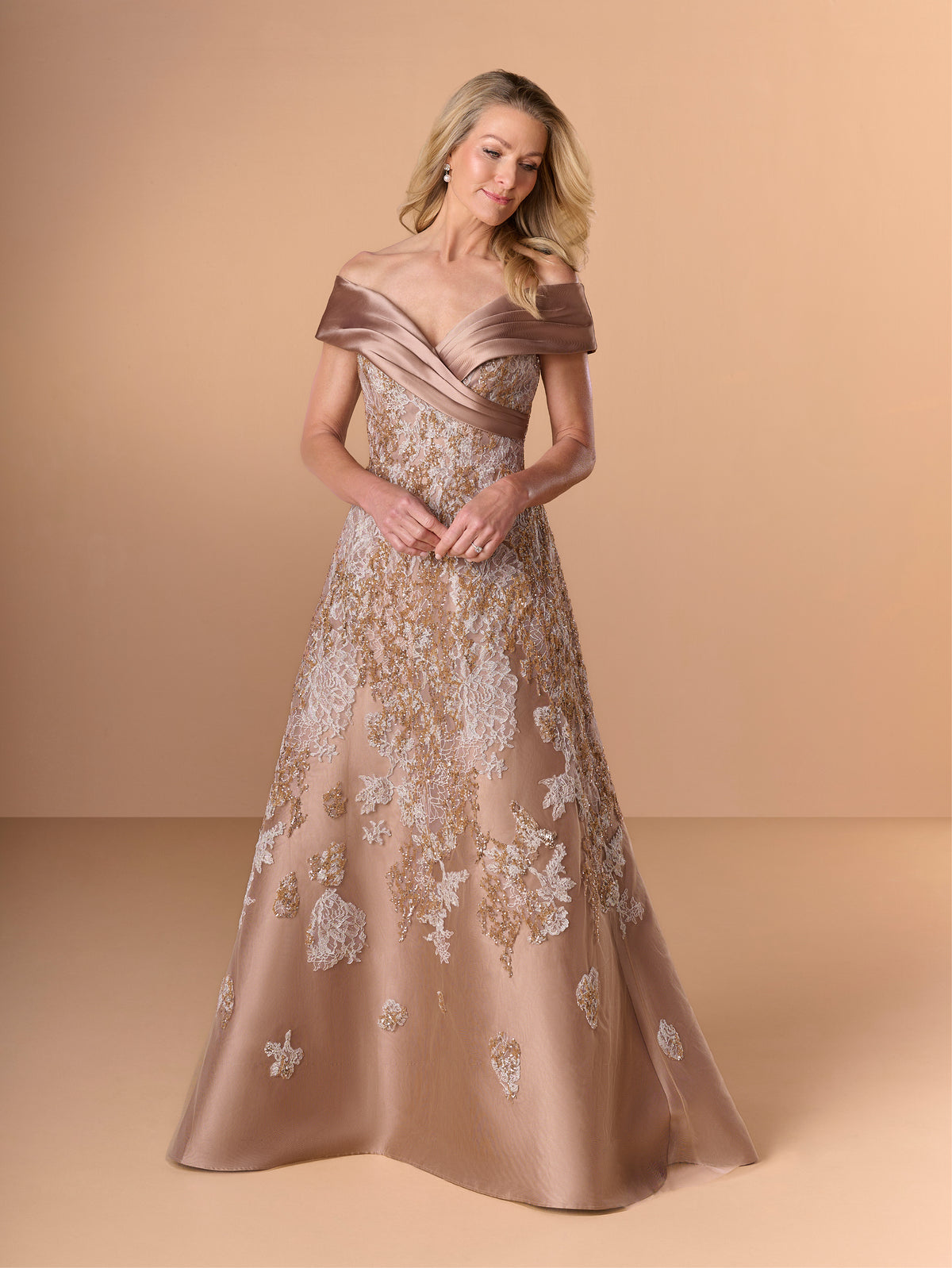 A woman models an off-the-shoulder A-line evening gown in a champagne beige satin fabric with white floral embroidery, set against a solid warm beige studio background. / A-line lace dress with mikado collar off-shoulder.