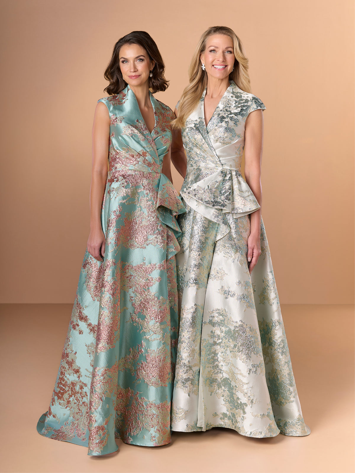 Two women modeling formal evening gowns against a warm beige studio backdrop. The gown on the left is sleeveless with a wrap-style bodice and a flowing skirt in aqua with pink floral patterning. / A-line style in brocade with V-neck and standing collar at the back neckline. Side front pleating on bodice finished with short ruffle.