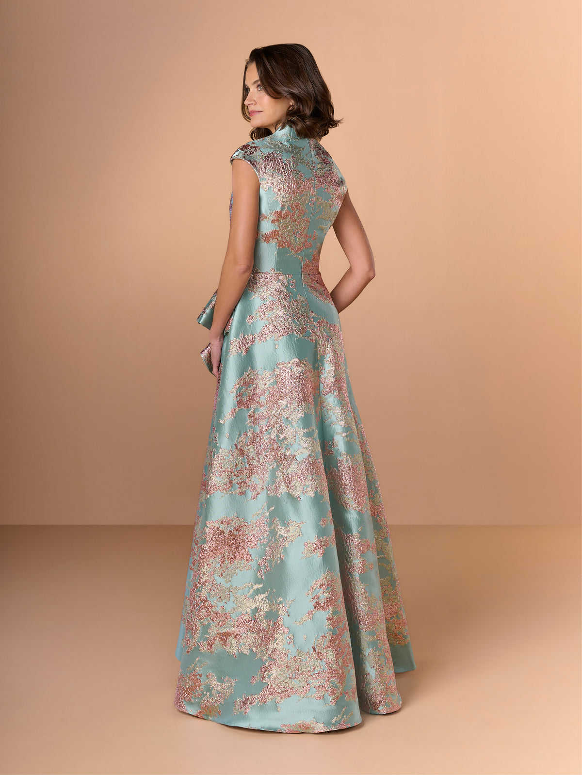Woman wearing a floral evening gown against a beige background / A-line style in brocade with V-neck and standing collar at the back neckline. Side front pleating on bodice finished with short ruffle.
