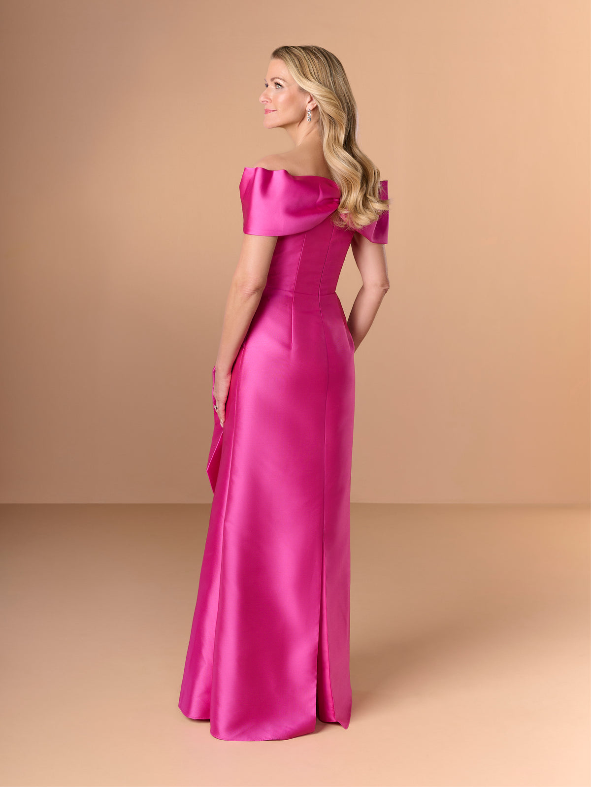 A woman modeling a bright fuchsia satin off-the-shoulder evening gown, shown from a side-back angle against a solid warm beige studio backdrop. / Slim mikado style with straight neckline and off-shoulder straps. Draping on bust area and frill feature on side front skirt.