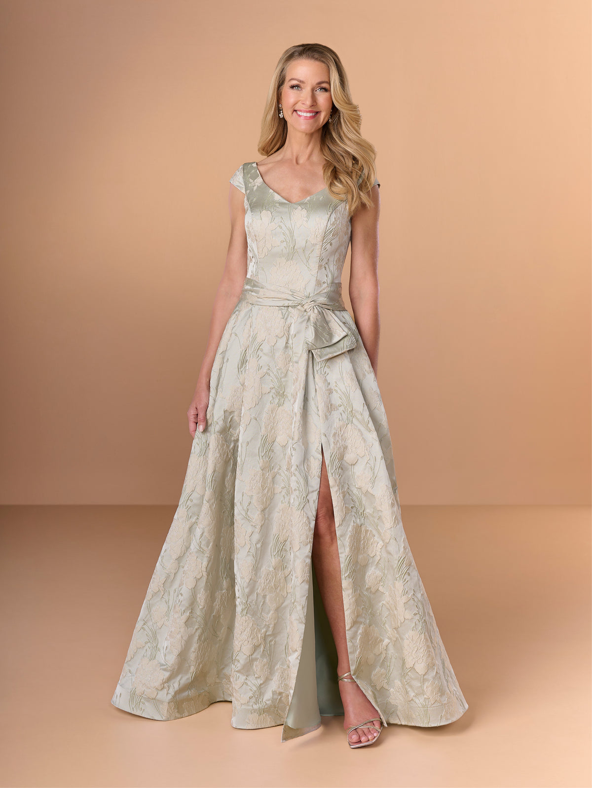 A woman modeling a sleeveless floor-length formal gown with a fitted bodice, wide flared skirt, and front slit, in a pale sage green and champagne brocade fabric against a warm beige studio backdrop. / A-line brocade style with V-neck and short sleeves. Waist band draping with soft bow on side. Invisible zipper closure at the back.