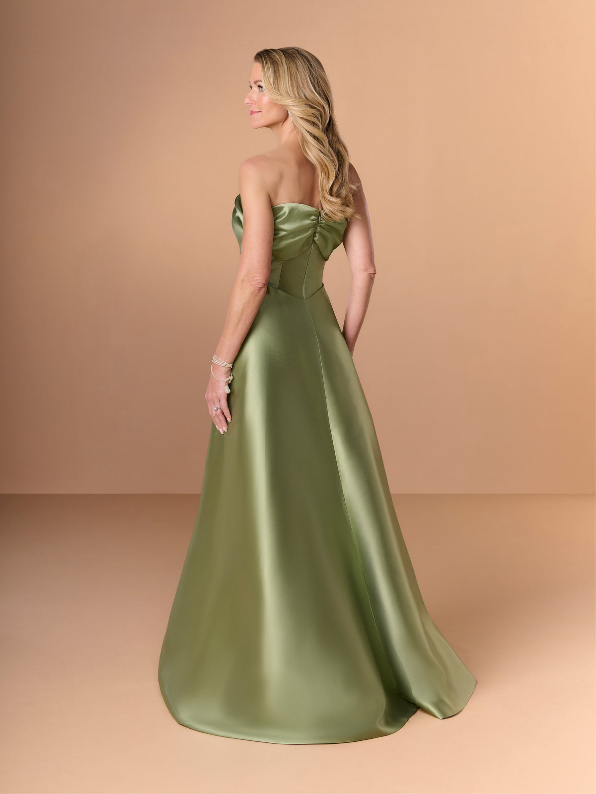 A woman modeling a strapless satin ball gown in sage green, shown from the back against a warm beige studio backdrop. / Slim mikado style with draping on bust area in bow shape going around to the back. Side front draping on stomach area. Detachable back skirt sweep.