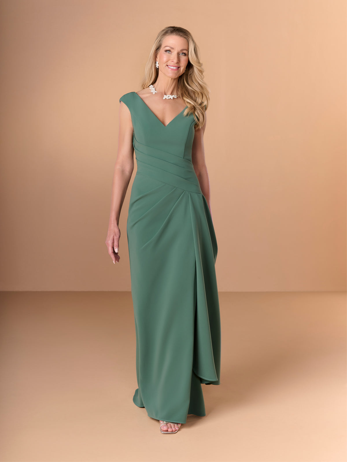 A woman modeling a floor-length sage green gown with a V-neckline, cap sleeves, and a draped gathered waist, photographed against a warm beige studio backdrop. / Slim crepe style with V-neck and cup shoulder. Side pleating on front dress with long ruffle on side.