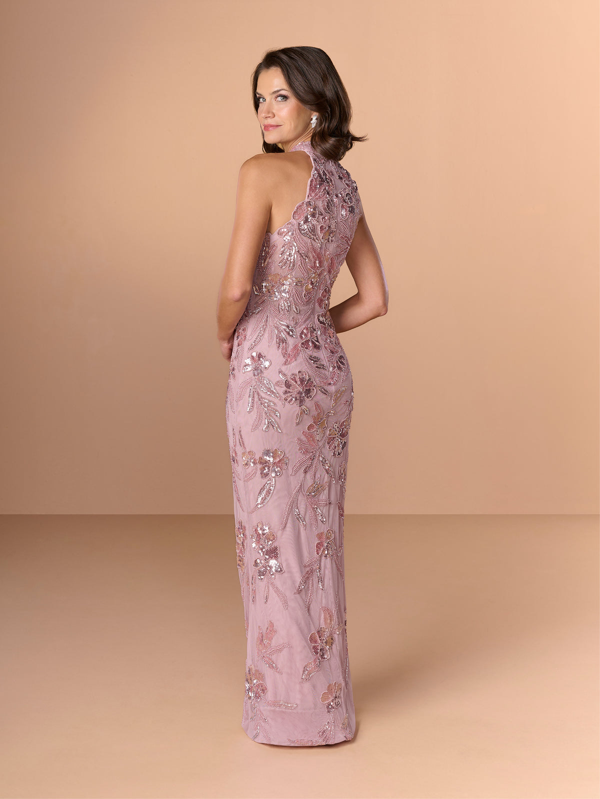 Woman wearing a pink floral evening gown against a beige background / This dress is a fitted, floor-length gown featuring a high halter neckline and an all-over floral pattern made of shimmering sequins and embroidery. The design follows a sleek, straight silhouette that highlights the embellished details from top to bottom, creating an elegant and refined look.