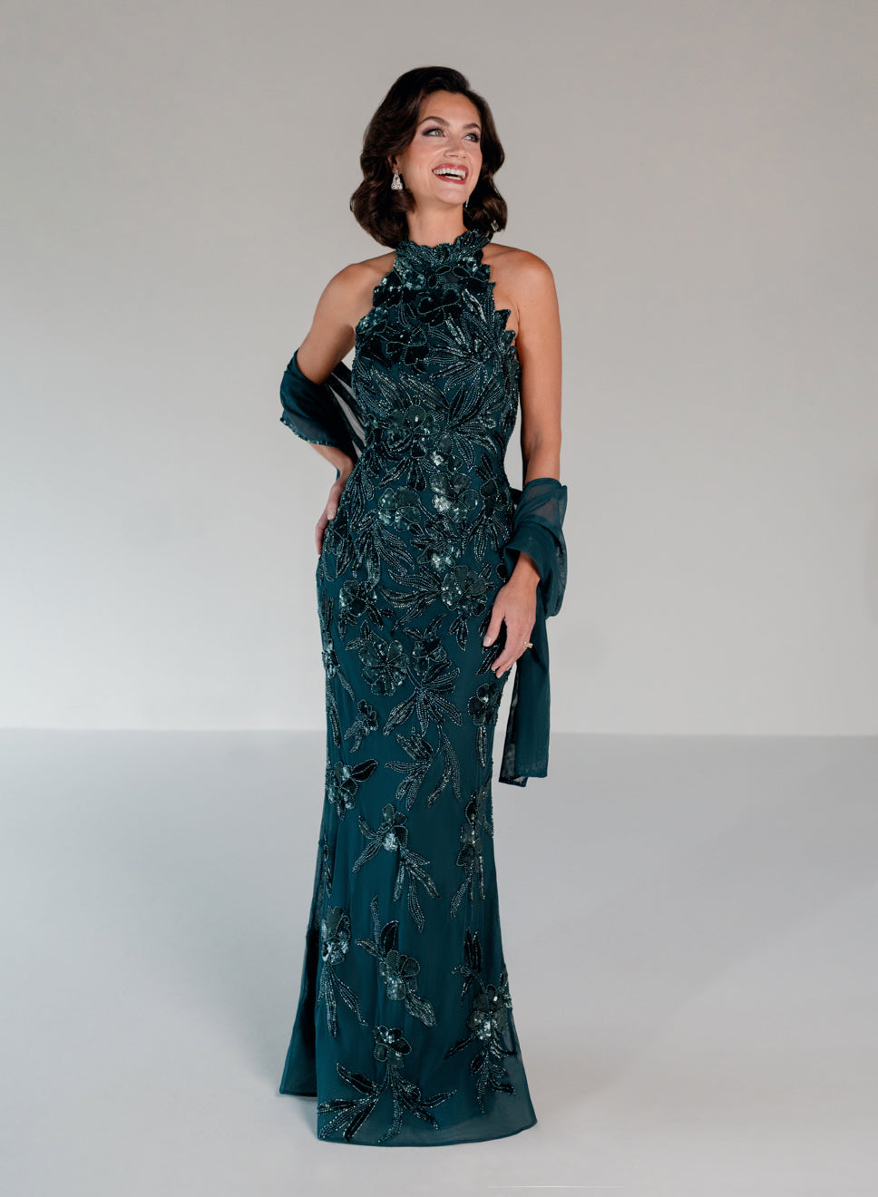 A woman modeling a floor-length dark teal evening gown with intricate floral beadwork and sequins, paired with matching sheer draped gloves, against a neutral light gray studio background. / This dress is a fitted, floor-length gown featuring a high halter neckline and an all-over floral pattern made of shimmering sequins and embroidery. The design follows a sleek, straight silhouette that highlights the embellished details from top to bottom, creating an elegant and refined look.