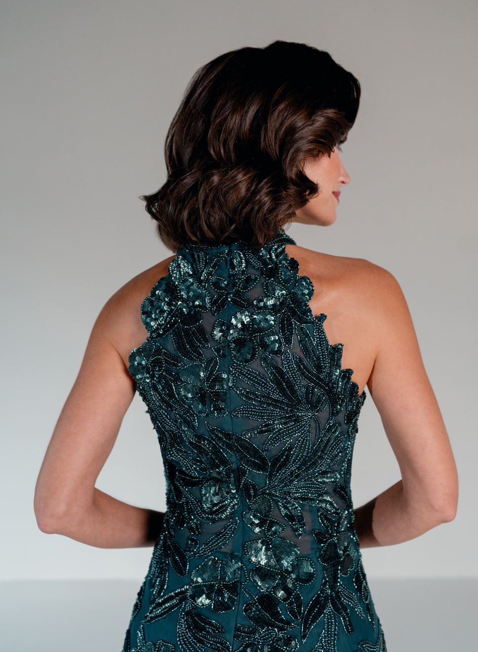 A woman shown from the back wearing a sleeveless halter-neck evening gown with intricate dark teal sequined floral embroidery over sheer mesh. / This dress is a fitted, floor-length gown featuring a high halter neckline and an all-over floral pattern made of shimmering sequins and embroidery. The design follows a sleek, straight silhouette that highlights the embellished details from top to bottom, creating an elegant and refined look.