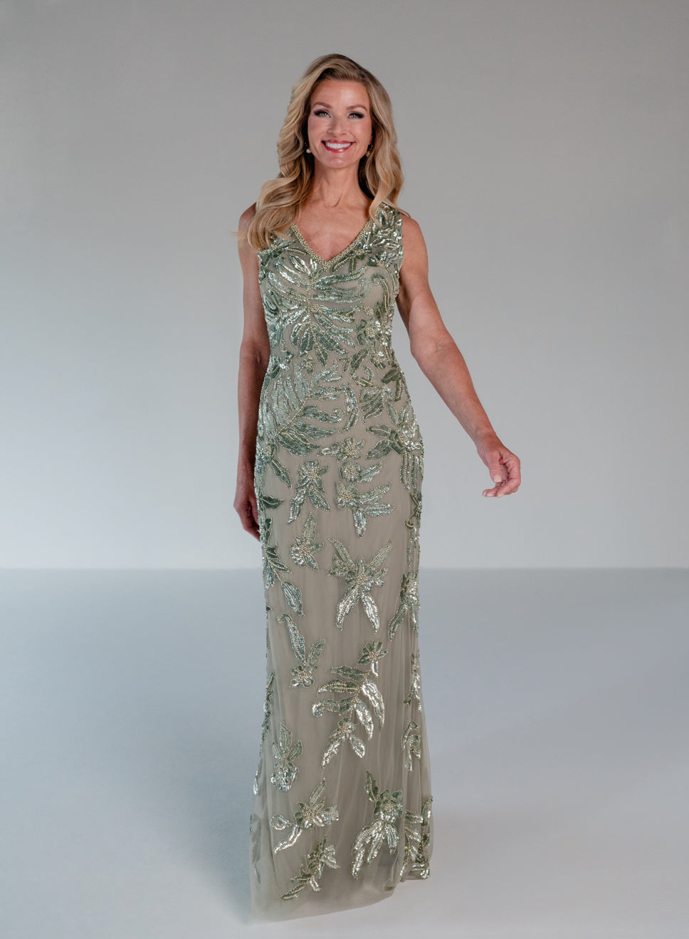 A woman modeling a sleeveless, floor-length evening gown with a V-neckline. The dress features a shimmering taupe-beige base covered in metallic silver and green leafy sequin embroidery, set against a plain light gray studio background. / This gown features a sleeveless, fitted silhouette with a softly draped v-neckline. The entire dress is decorated with shimmering floral and leaf-patterned embellishments that extend from top to hem. Its sleek, floor-length design creates an elegant and refined look from b