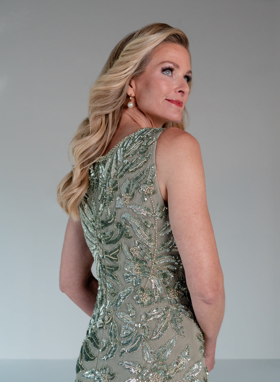 A blonde woman wearing a sleeveless, one-shoulder embellished dress in a shimmering sage green and silver sequin pattern, photographed against a neutral gray studio background. / This gown features a sleeveless, fitted silhouette with a softly draped v-neckline. The entire dress is decorated with shimmering floral and leaf-patterned embellishments that extend from top to hem. Its sleek, floor-length design creates an elegant and refined look from both the front and back.