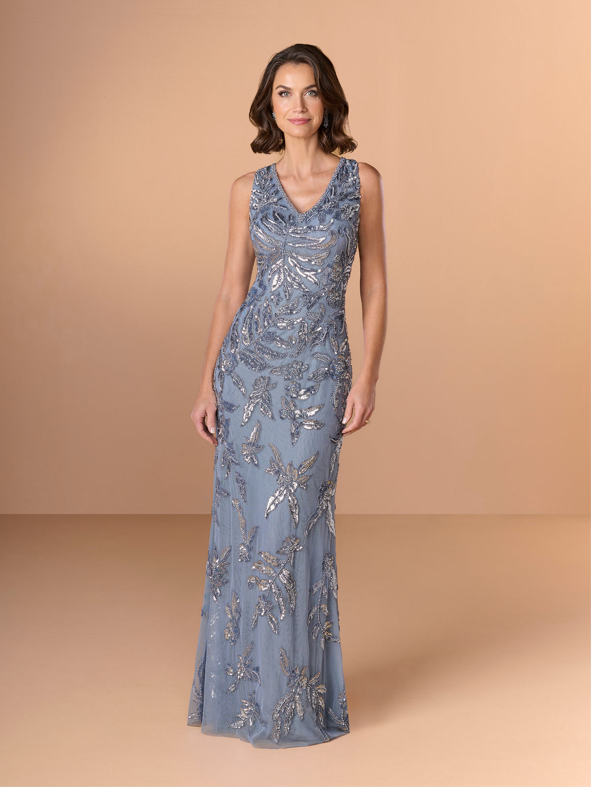 Woman wearing a long, patterned dress against a beige background / This gown features a sleeveless, fitted silhouette with a softly draped v-neckline. The entire dress is decorated with shimmering floral and leaf-patterned embellishments that extend from top to hem. Its sleek, floor-length design creates an elegant and refined look from both the front and back.
