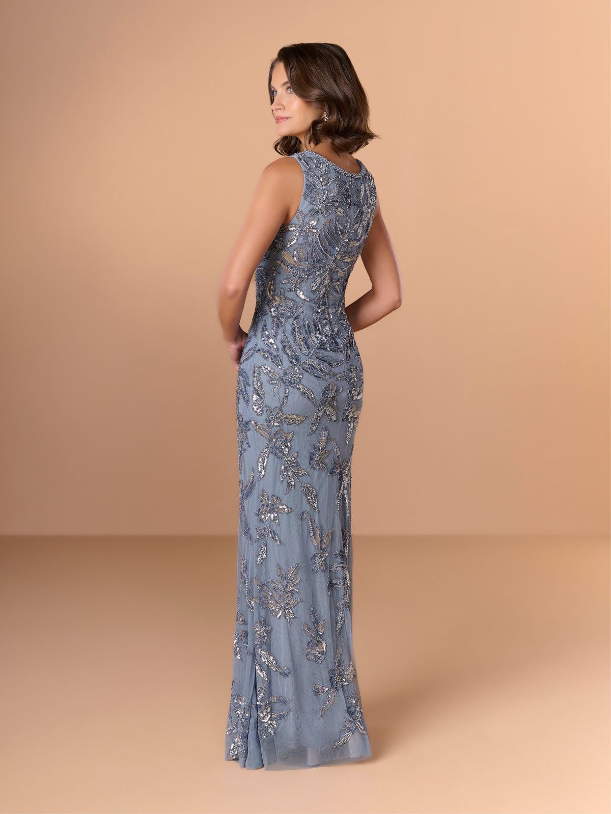A woman modeling a sleeveless floor-length gown from the back, featuring intricate silver beaded floral embroidery over a dusty blue-gray base, set against a solid warm beige studio backdrop. / This gown features a sleeveless, fitted silhouette with a softly draped v-neckline. The entire dress is decorated with shimmering floral and leaf-patterned embellishments that extend from top to hem. Its sleek, floor-length design creates an elegant and refined look from both the front and back.