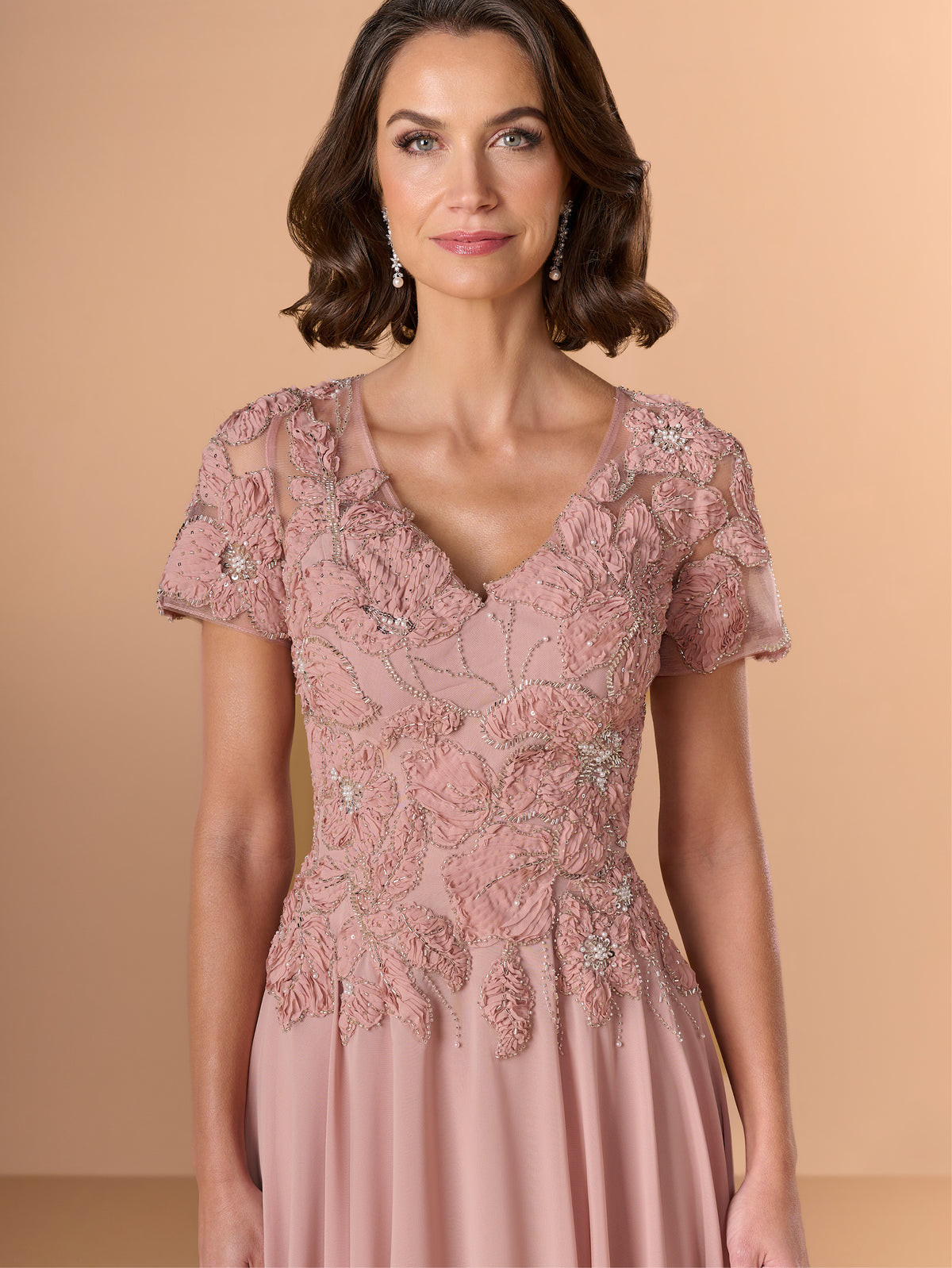 A woman wearing a dusty pink evening gown with intricate floral lace embroidery, short sleeves, and a V-neckline, set against a solid beige studio background. / A-line chiffon style with bodice coved with beaded motifs, V-neck short semi sheer sleeves.