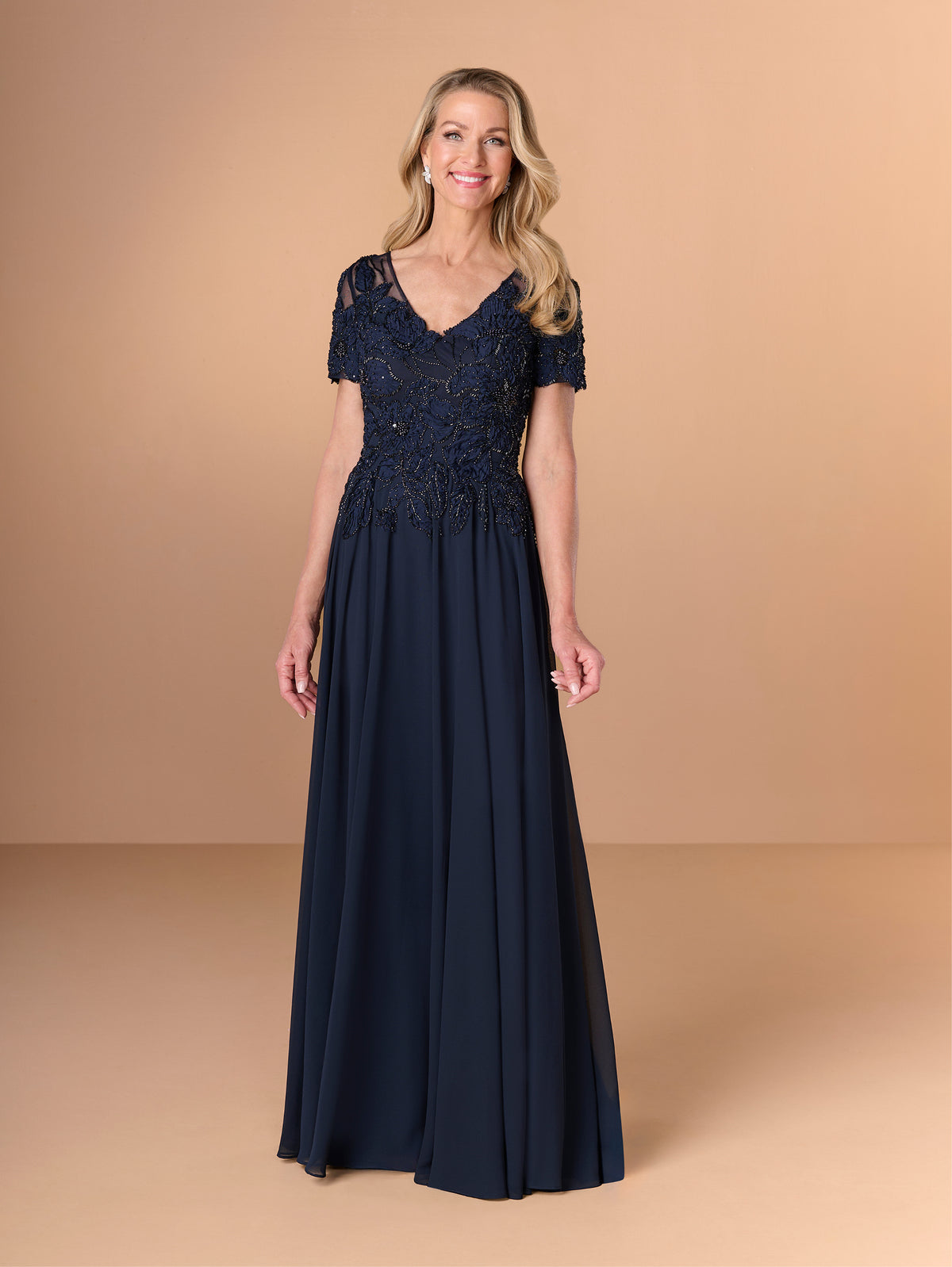 A woman modeling a floor-length navy blue gown with a lace bodice, short lace sleeves, and a flowing chiffon skirt against a solid beige studio background. / A-line chiffon style with bodice coved with beaded motifs, V-neck short semi sheer sleeves.