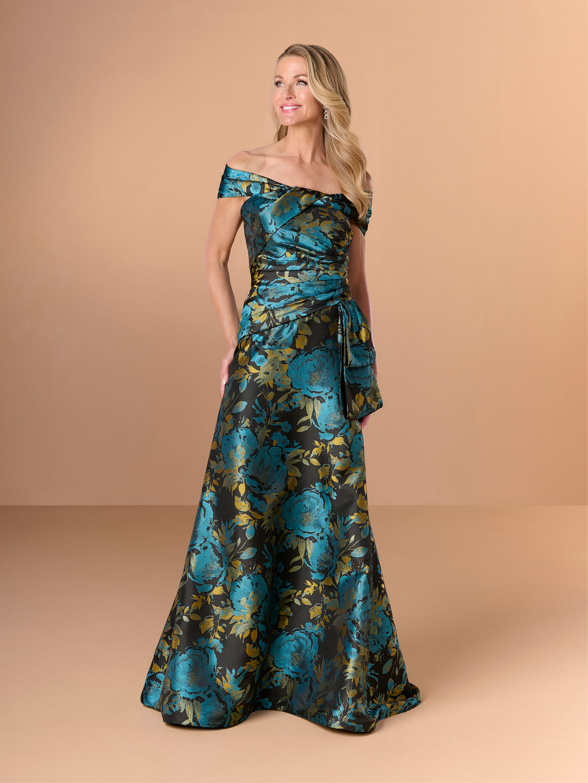 A woman modeling an off-the-shoulder floor-length gown with a teal and black floral jacquard pattern, set against a solid beige studio background. / Slim brocade style with collar/straps twist, pleating on front bodice.
