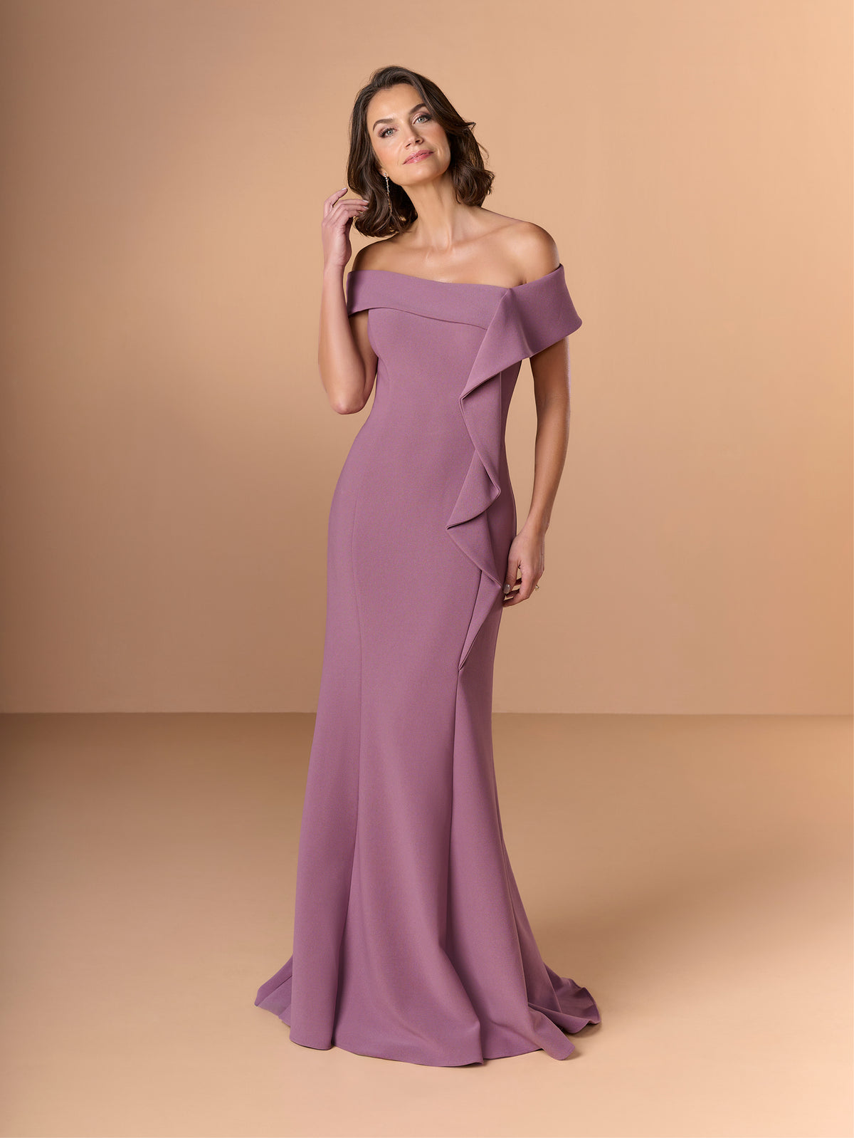 A woman modeling an off-the-shoulder floor-length gown in a dusty mauve-purple color, set against a solid warm beige studio background. / Slim crepe style with asymmetrical wide collar and side raffle.