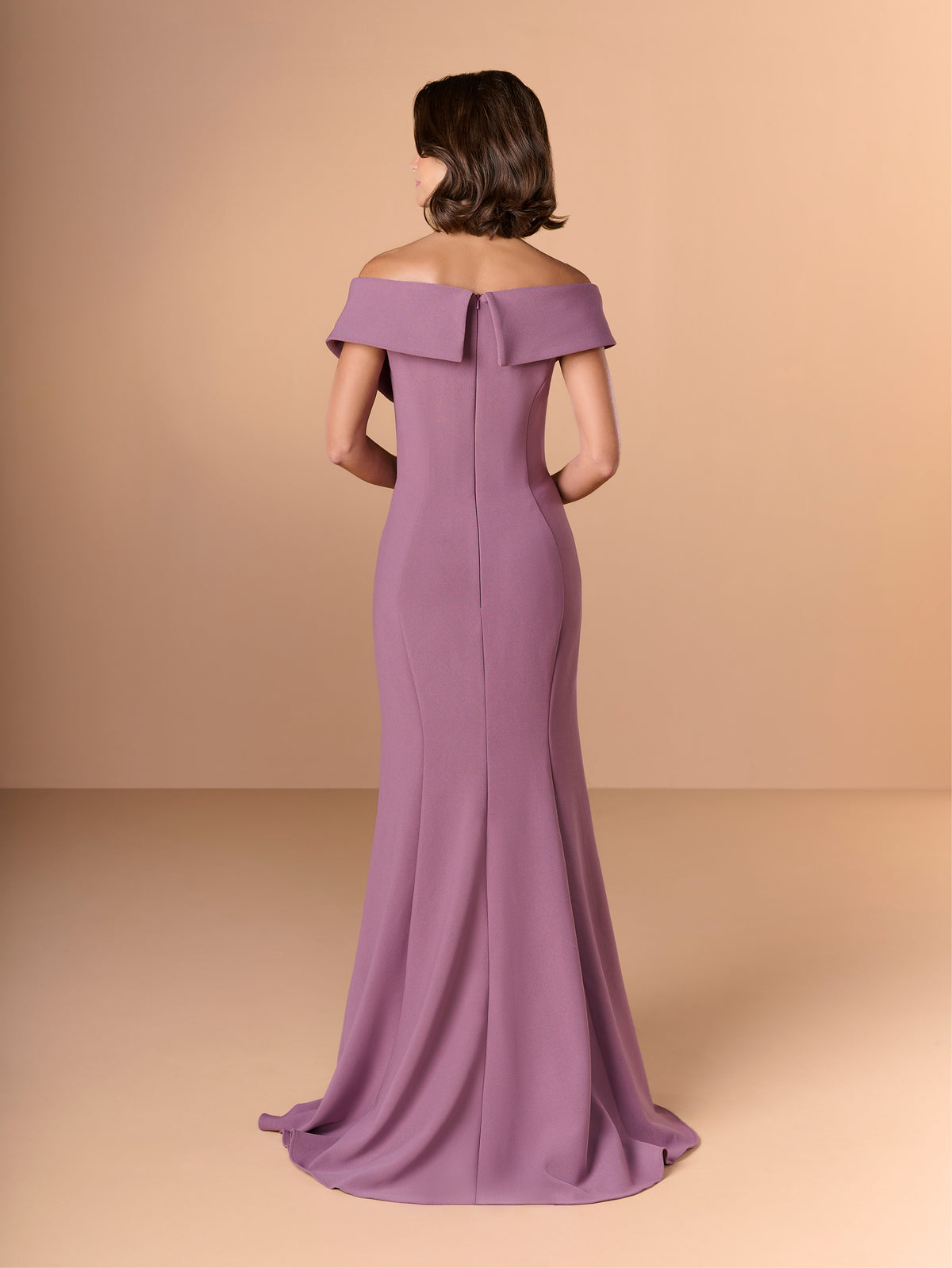 A woman modeling an off-the-shoulder floor-length gown from the back, shown against a warm beige studio backdrop. The dress is a muted dusty lavender-purple with a structured bow detail at the back neckline and a slight train. / Slim crepe style with asymmetrical wide collar and side raffle.