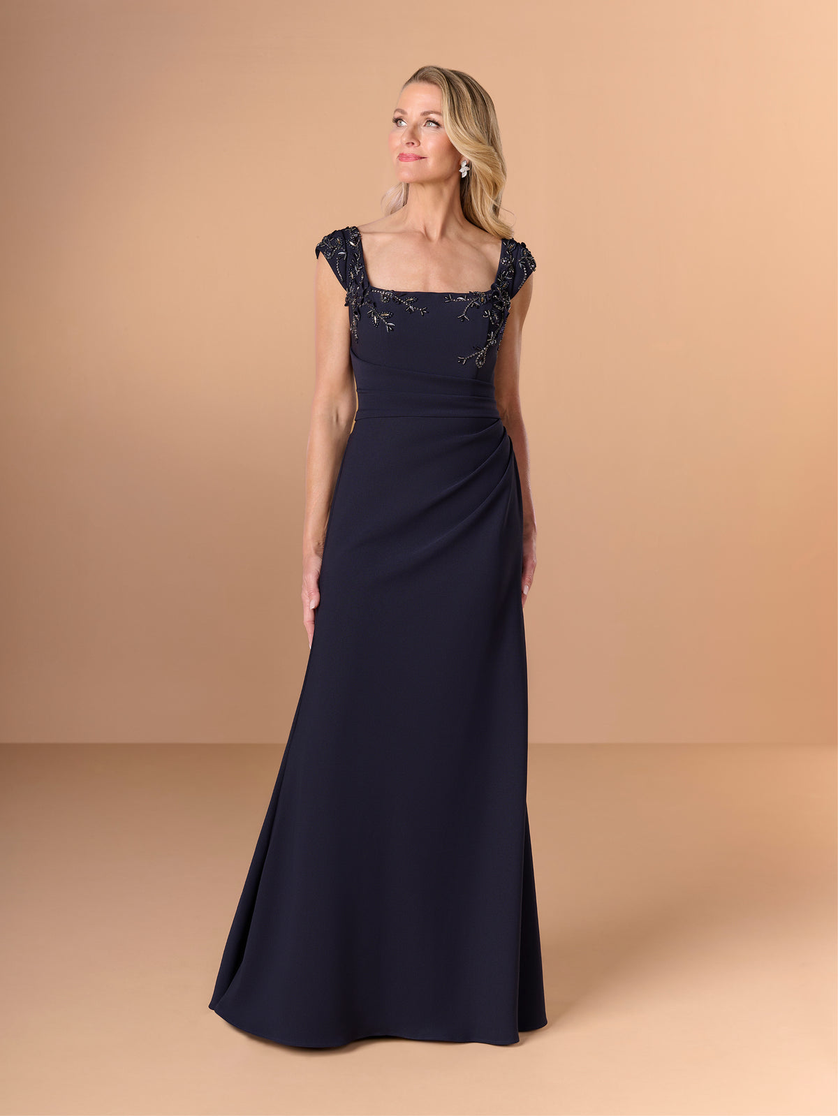 A woman modeling a floor-length navy evening gown with cap sleeves, a square neckline, and lace embellishment across the bodice, set against a solid warm beige studio background. / Cap sleeve fitted gown with pleated detail on the bodice and 3D embellishment.