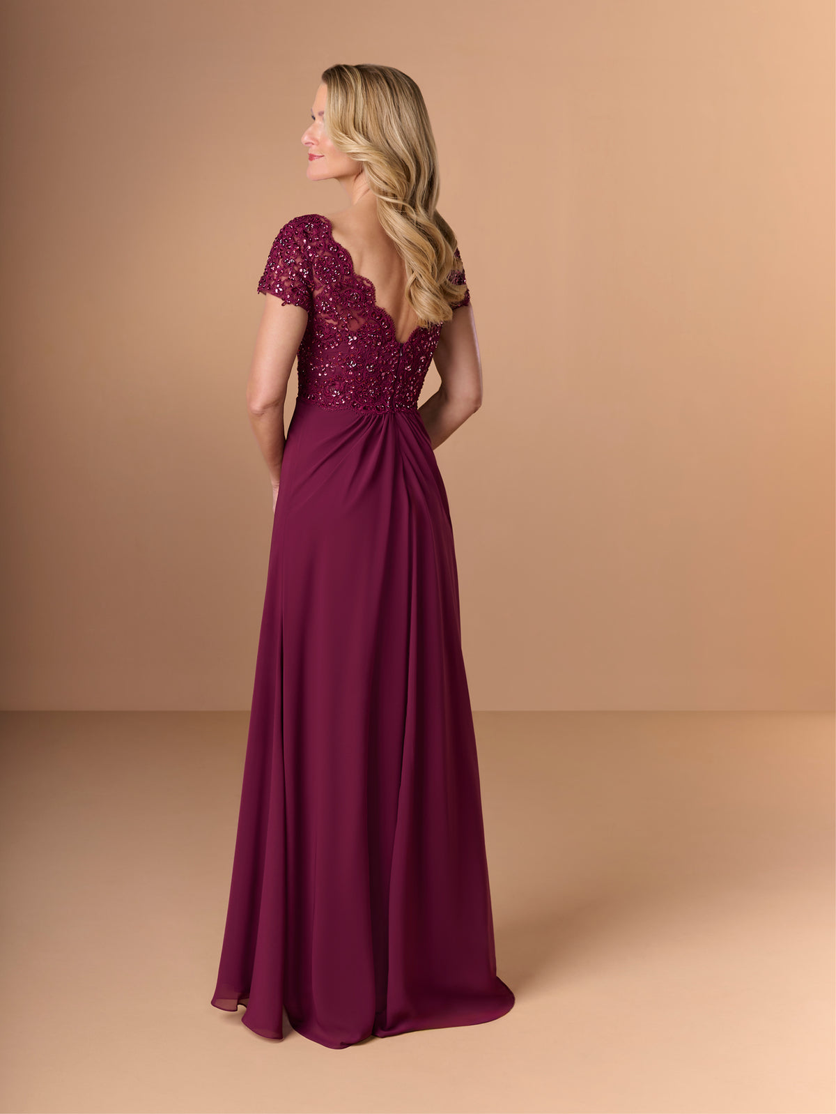A woman modeling a floor-length burgundy evening gown from the back, featuring a beaded lace bodice, short cap sleeves, and a flowing skirt, set against a solid beige studio background. / A-line style with lace bodice and chiffon skirt. V-neck edge and short sleeves hemline edge finished with scallops. Draping on front and back skirt in fan shape.