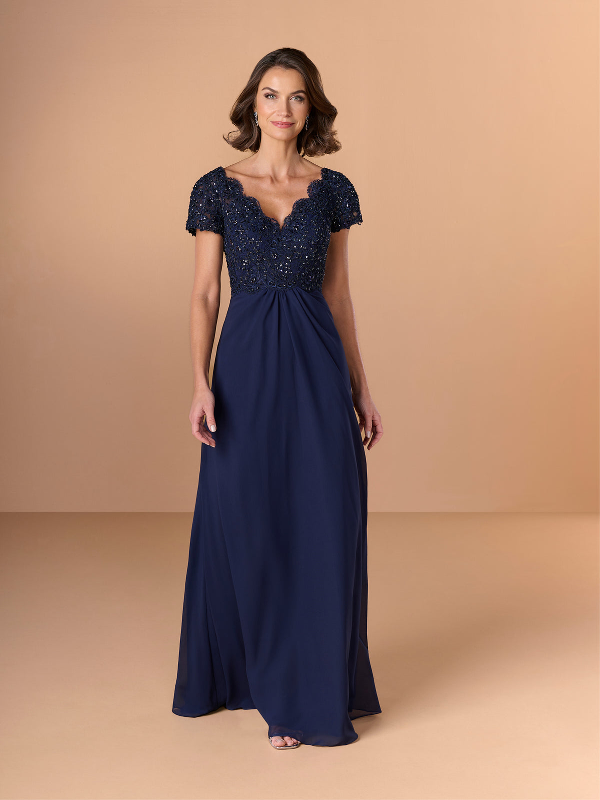 A woman modeling a navy blue floor-length gown with a beaded, scalloped sweetheart bodice and short cap sleeves, set against a solid beige studio backdrop. / A-line style with lace bodice and chiffon skirt. V-neck edge and short sleeves hemline edge finished with scallops. Draping on front and back skirt in fan shape.