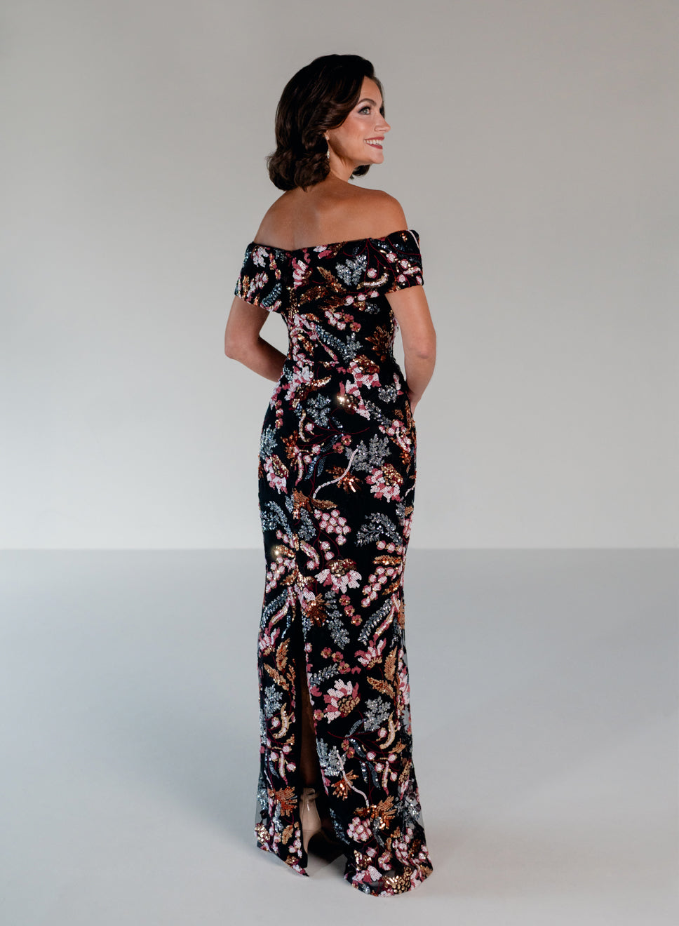 A woman modeling an off-the-shoulder black maxi dress with a vibrant pink, orange, and green floral and bird pattern, photographed against a neutral light gray studio background. / Slim lace style with straight neckline and wide off shoulder collar. Split on center back skirt.