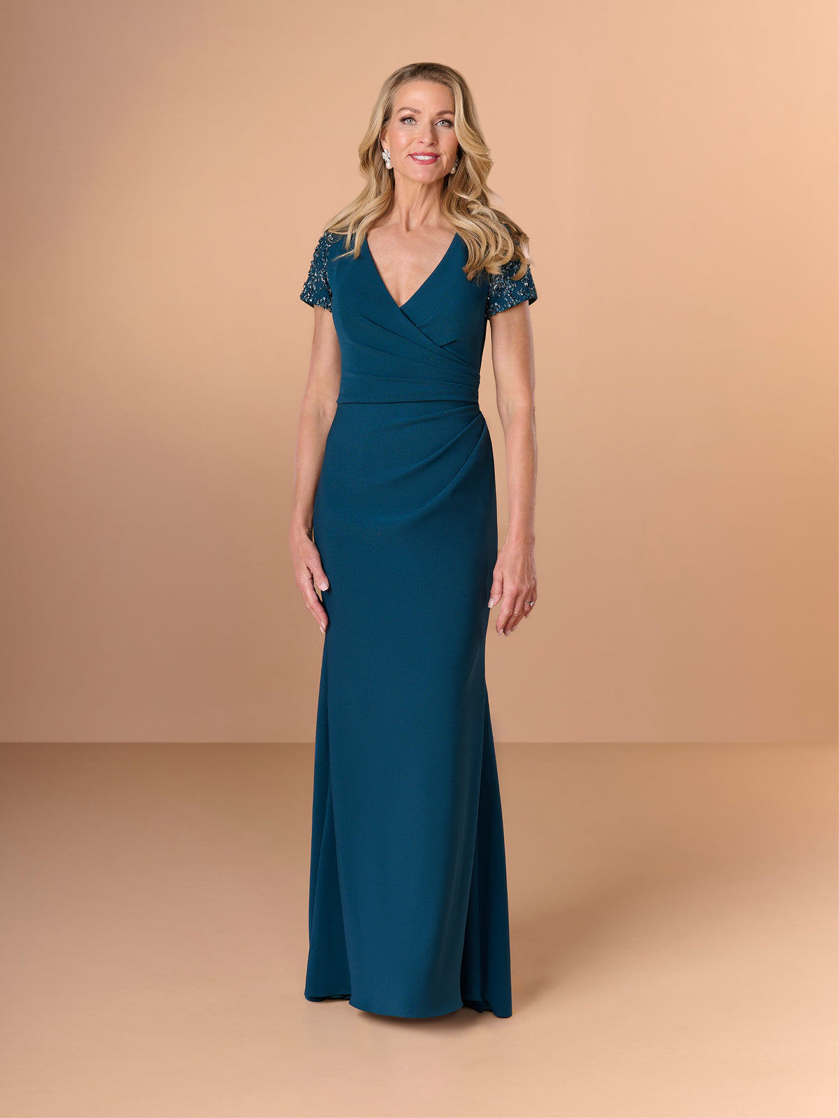 Woman wearing a teal evening gown with lace sleeves on a beige background / Short sleeve gown with beaded sleeve and body pleating detail.
