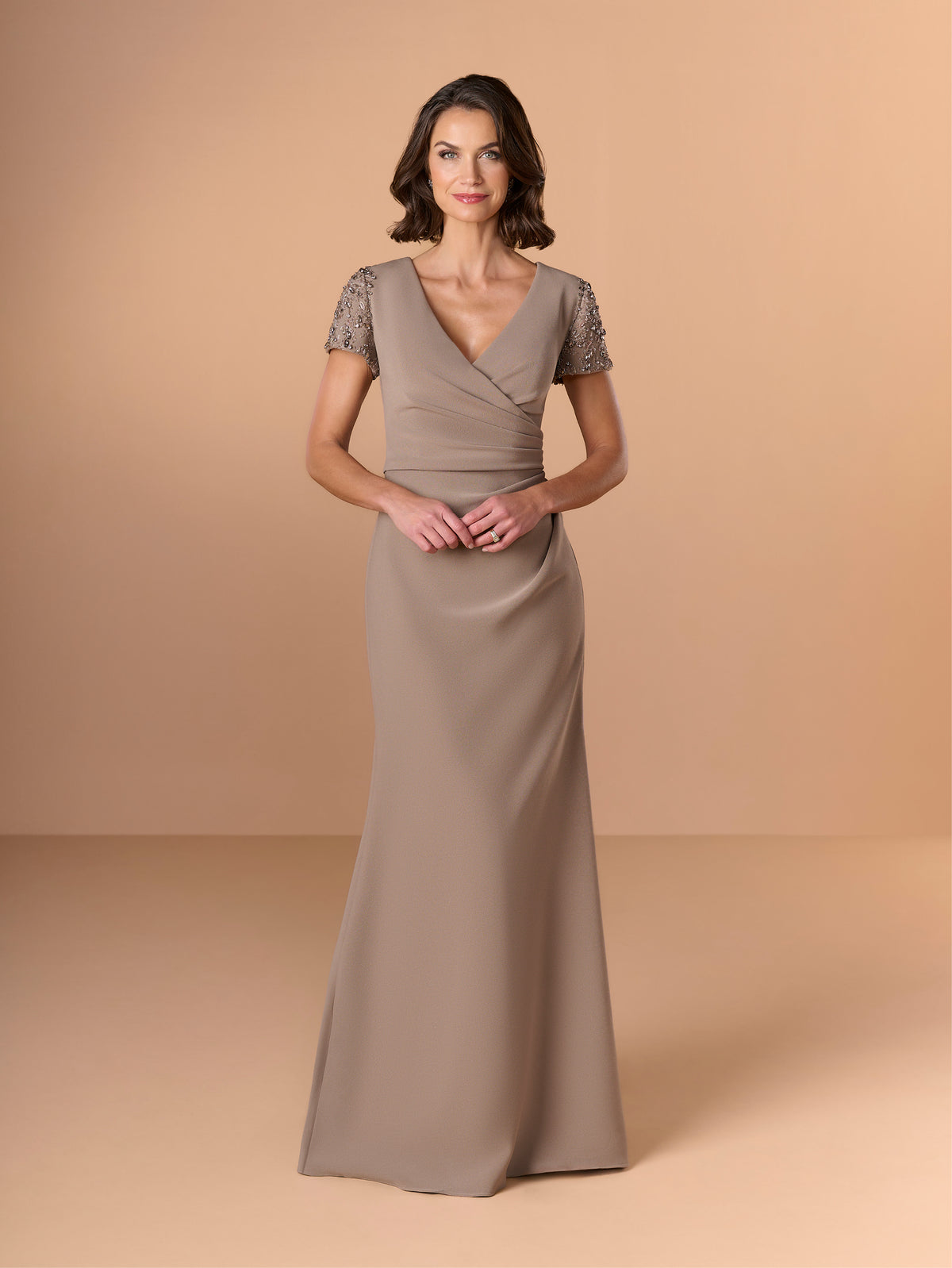 A woman modeling a floor-length taupe gown with lace short sleeves, a V-neckline, and a draped wrap-style bodice, photographed against a warm beige studio backdrop. / Short sleeve gown with beaded sleeve and body pleating detail.