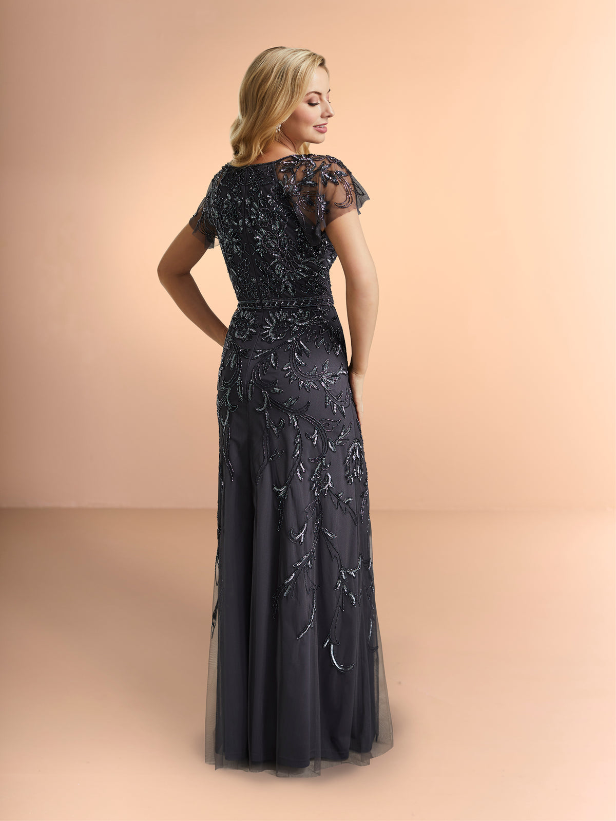 Woman wearing a dark blue evening gown with floral embroidery on a beige background / Slim long beaded style in soft mesh. Rounded neck and sheer short ruffle sleeves. Zipper back closure.