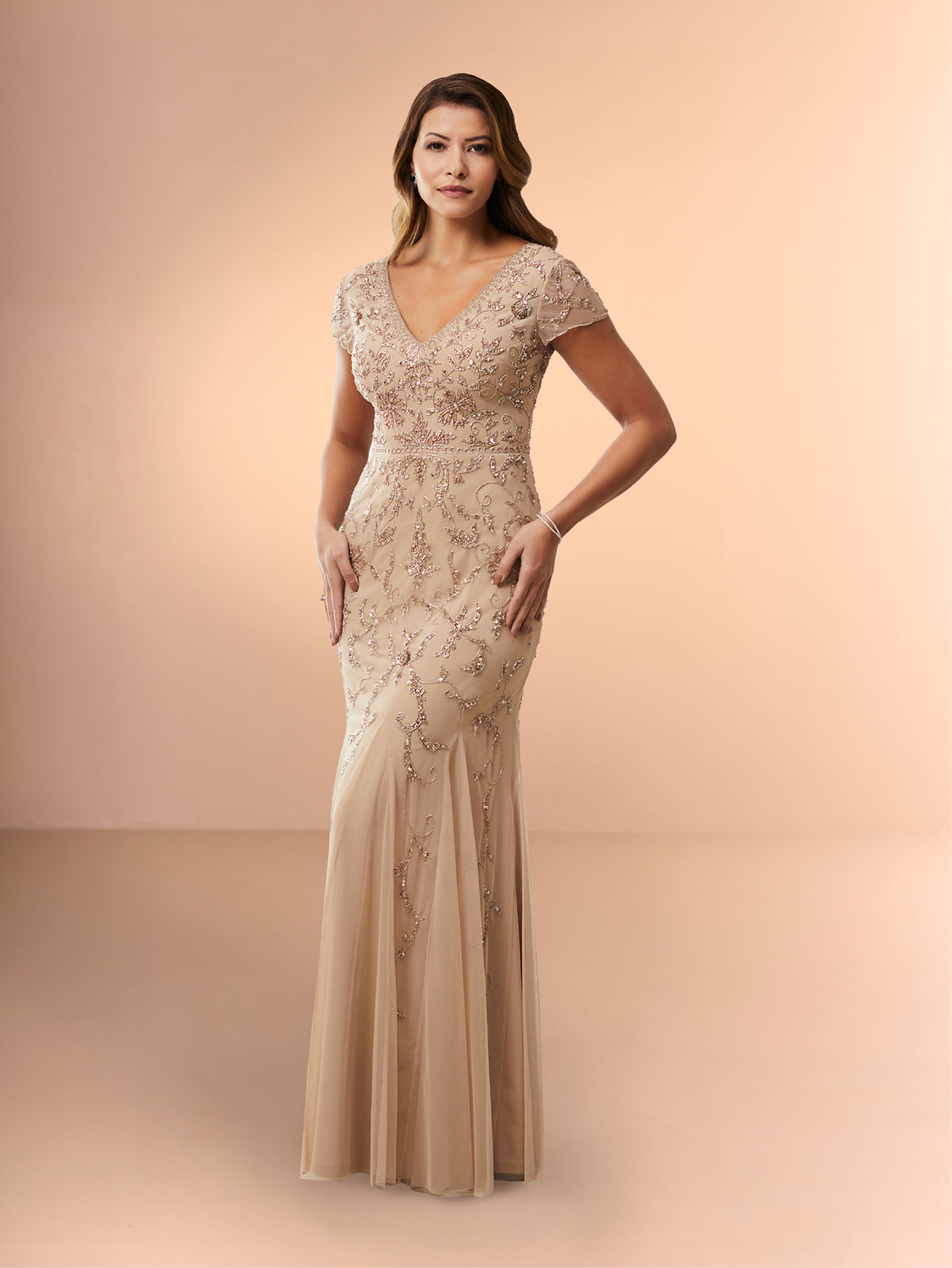 A woman modeling a floor-length champagne beige gown with intricate beaded floral embroidery and a V-neckline, set against a solid warm beige studio backdrop. / Slim long beaded style in soft mesh. V-neck and sheer short sleeves. Zipper back closure.