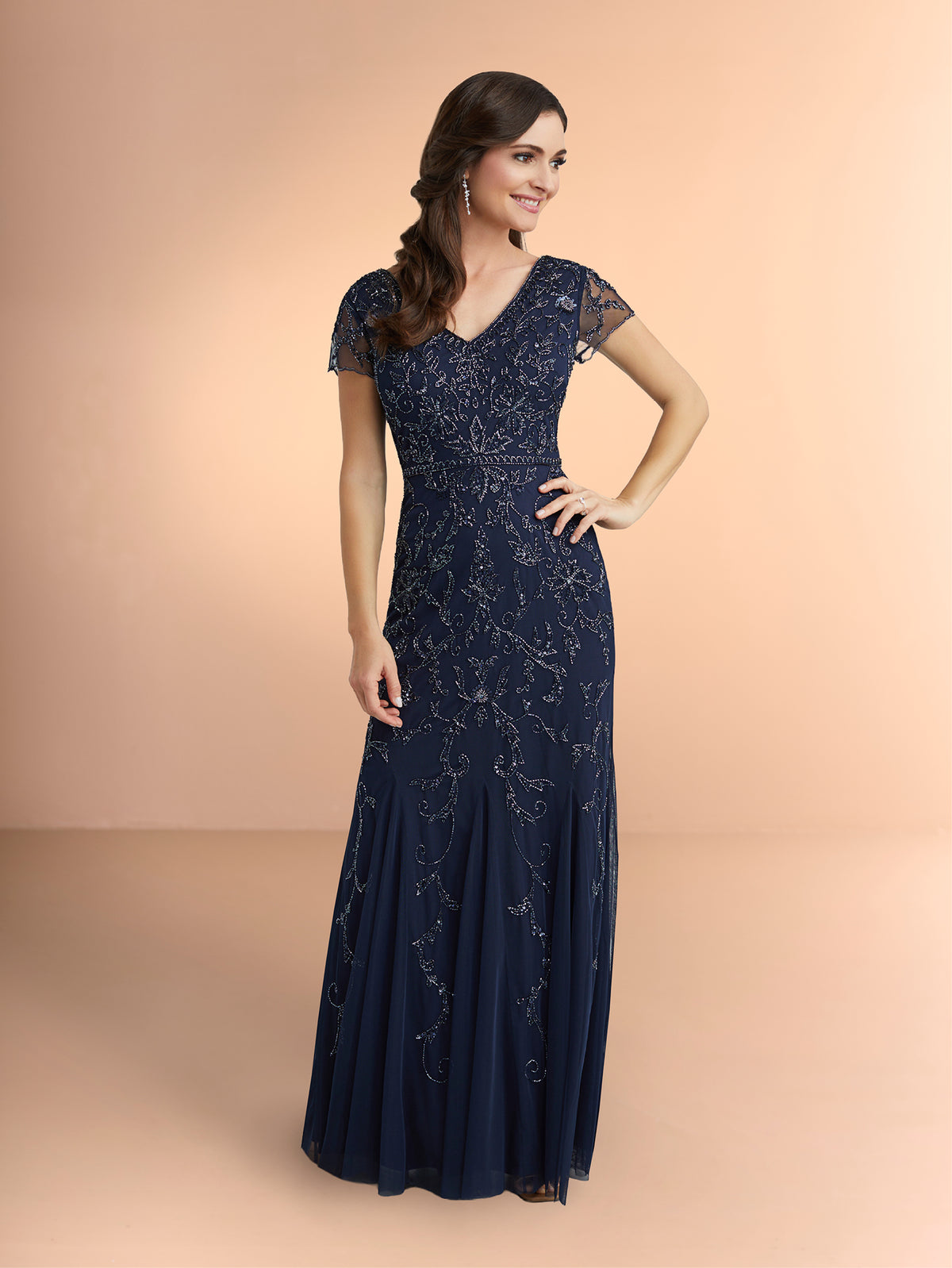 Woman wearing a navy blue lace evening gown on a peach background / Slim long beaded style in soft mesh. V-neck and sheer short sleeves. Zipper back closure.
