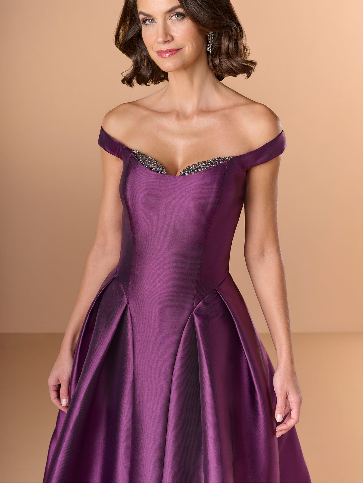 Woman wearing a purple off-shoulder gown with beaded details on a beige background / Off the shoulder gown with side skirt panels and inserted neckline beading.