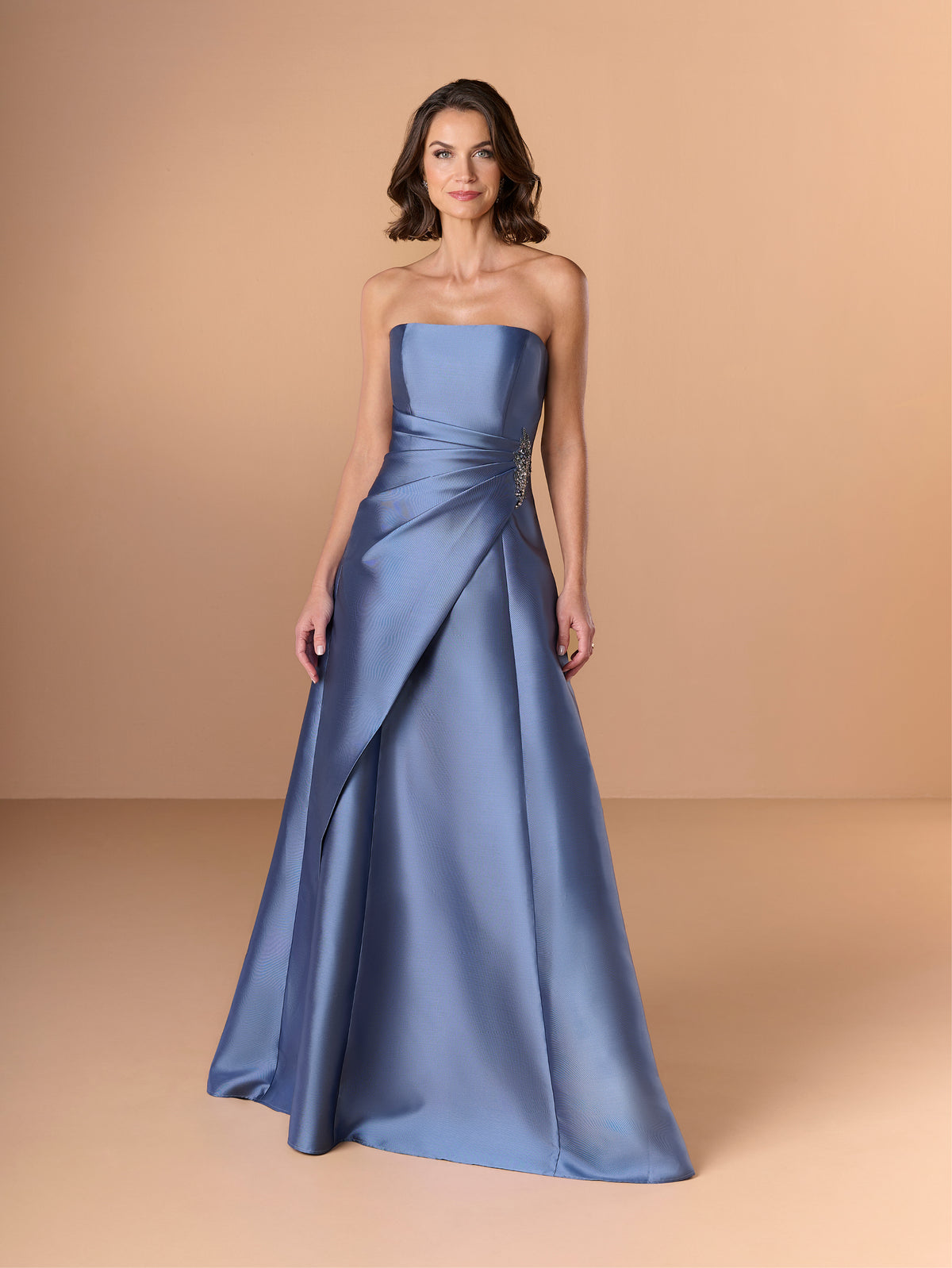 Woman wearing a strapless blue gown against a beige background / This strapless gown features a sleek, structured bodice that flows into an elegant A-line skirt. The design includes a dramatic, sculpted overlay at the waist, accented with a touch of sparkling embellishment on one side. Smooth, lustrous fabric and clean lines give the dress a sophisticated and modern look.