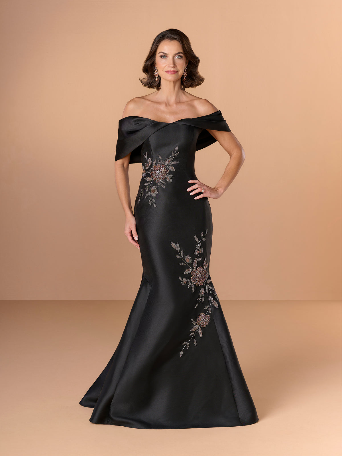A woman models an off-the-shoulder black satin evening gown with floral embroidery on a warm beige studio background. / Off the shoulder mermaid gown with placed beading.