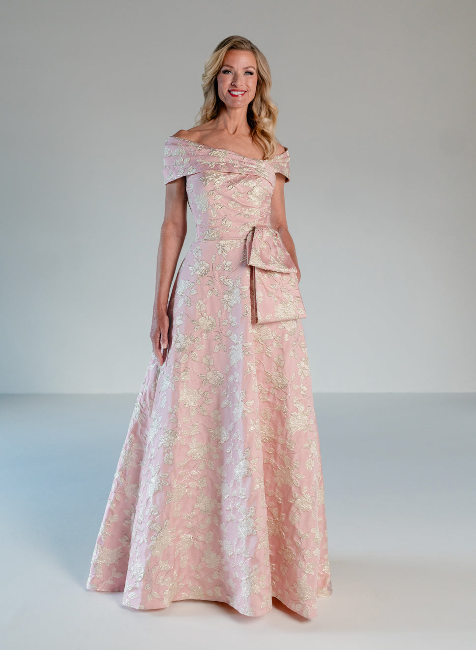 A woman modeling a floor-length off-the-shoulder gown in light pink with a subtle floral jacquard pattern and a matching bow detail at the waist, photographed against a neutral light gray studio background. / Brocade A-line style with off-shoulder collar/straps. Side front pleating on bodice finished with bow on side front waist area.
