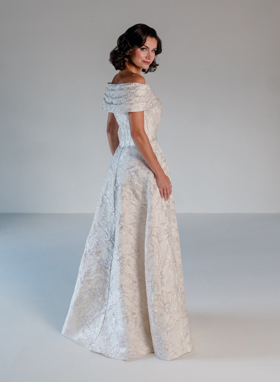 A woman modeling an off-the-shoulder floor-length gown with a fitted bodice and full skirt, photographed in a studio against a light gray background. / Brocade A-line style with off-shoulder collar/straps. Side front pleating on bodice finished with bow on side front waist area.