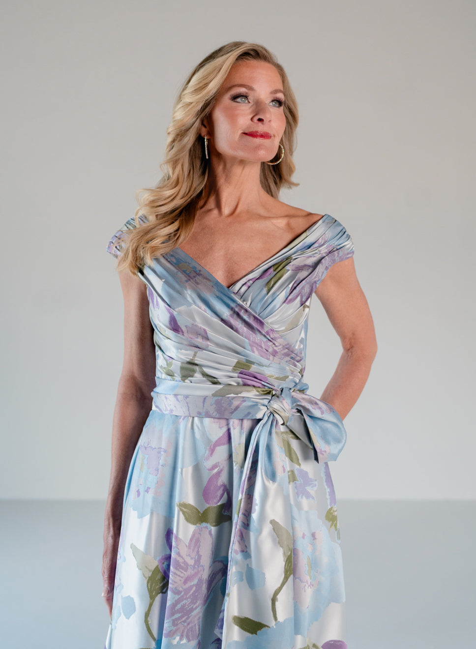 A woman modeling a light blue and white floral wrap dress with purple and green accents, set against a plain light gray studio background. / A-line style in jacquard. Pleated front bodice with V-neck and trim with bow on left side front waist area.